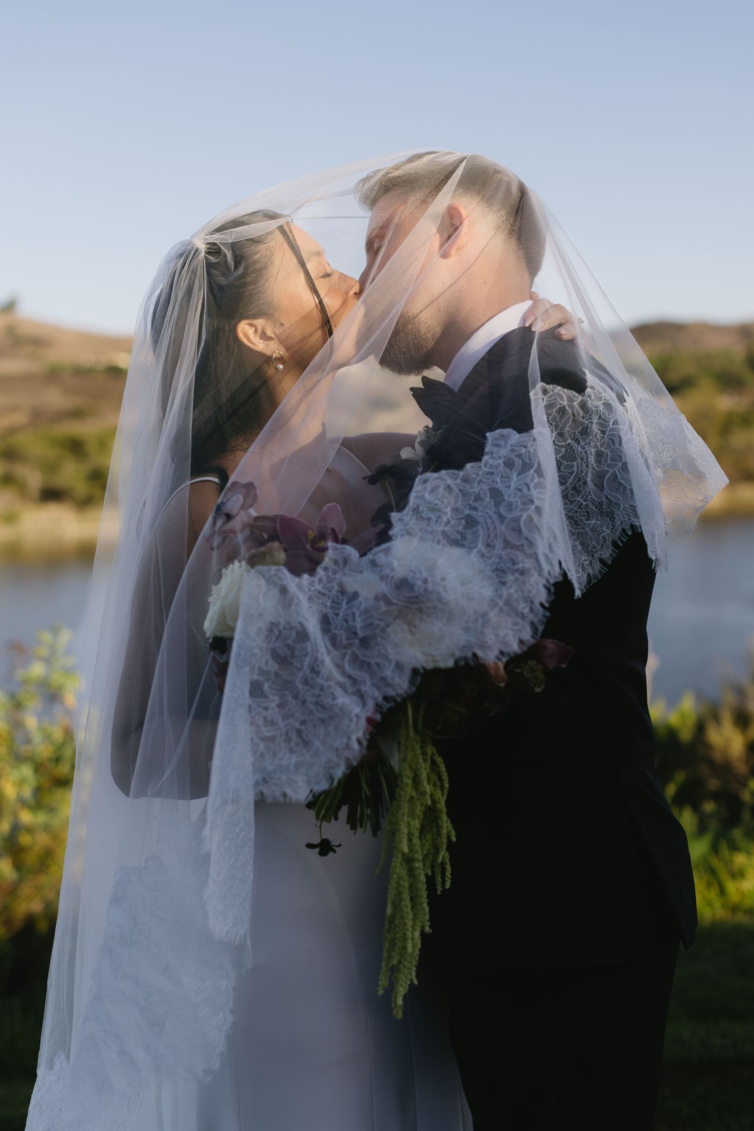 Romantic silhouette of newlyweds kissing by a lake at sunset with bride's veil flowing.