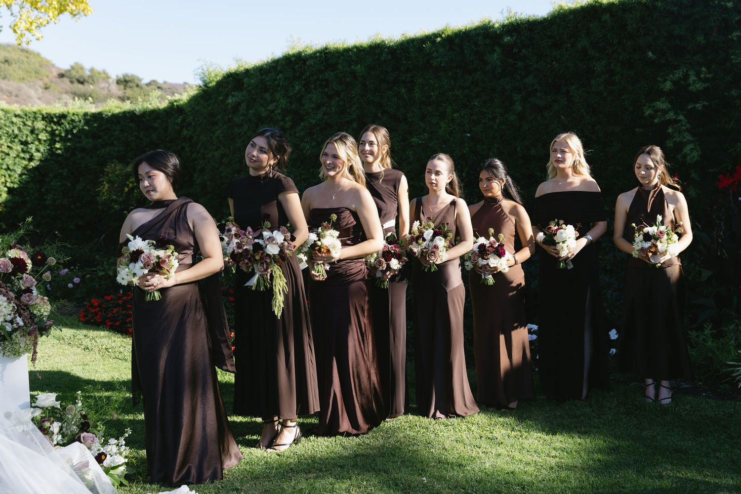 Bridesmaids in elegant brown dresses stand in a row holding floral bouquets against a hedge backdrop.