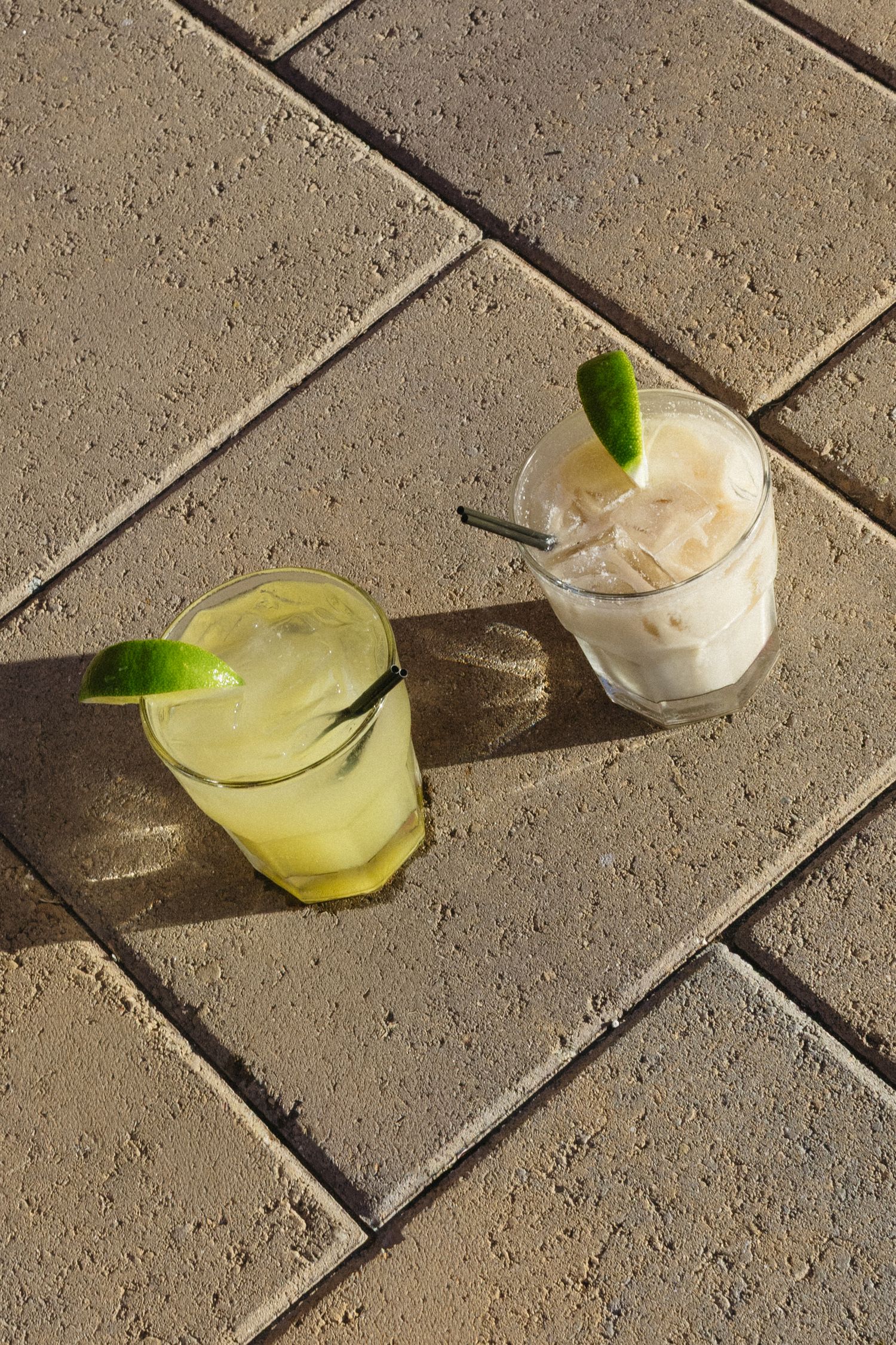 Refreshing cocktails with lime garnish served on stone tile surface in sunlight.