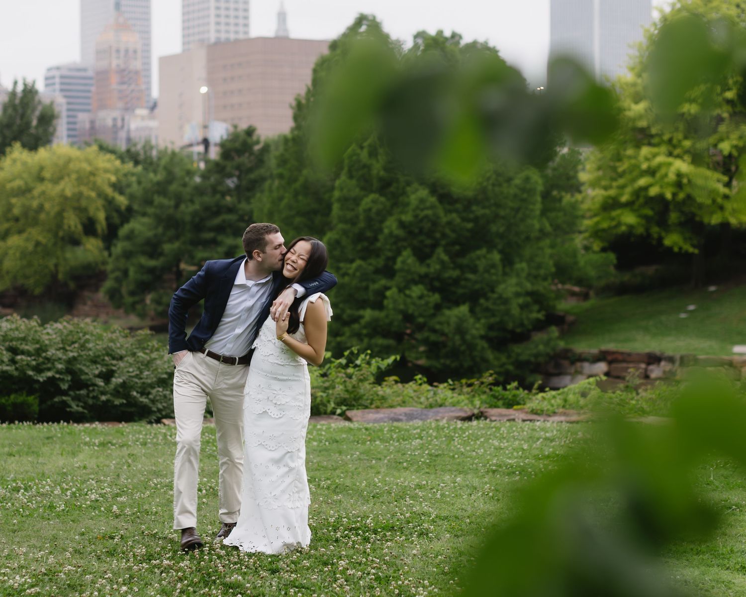 A couple poses against a city skyline backdrop with green foliage in the foreground.