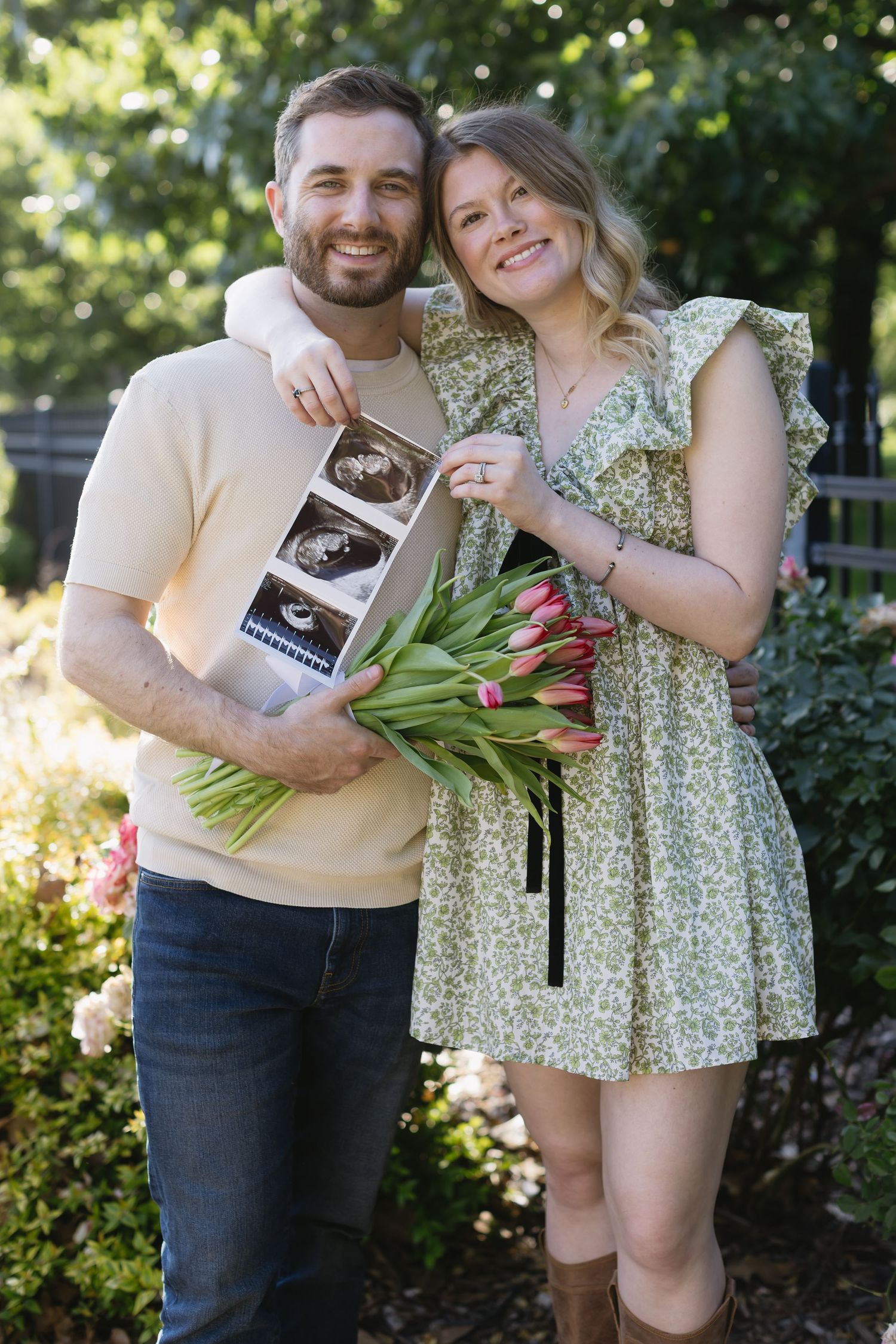 A couple poses with tulips and ultrasound photos in a garden during a springtime pregnancy announcement photoshoot.
