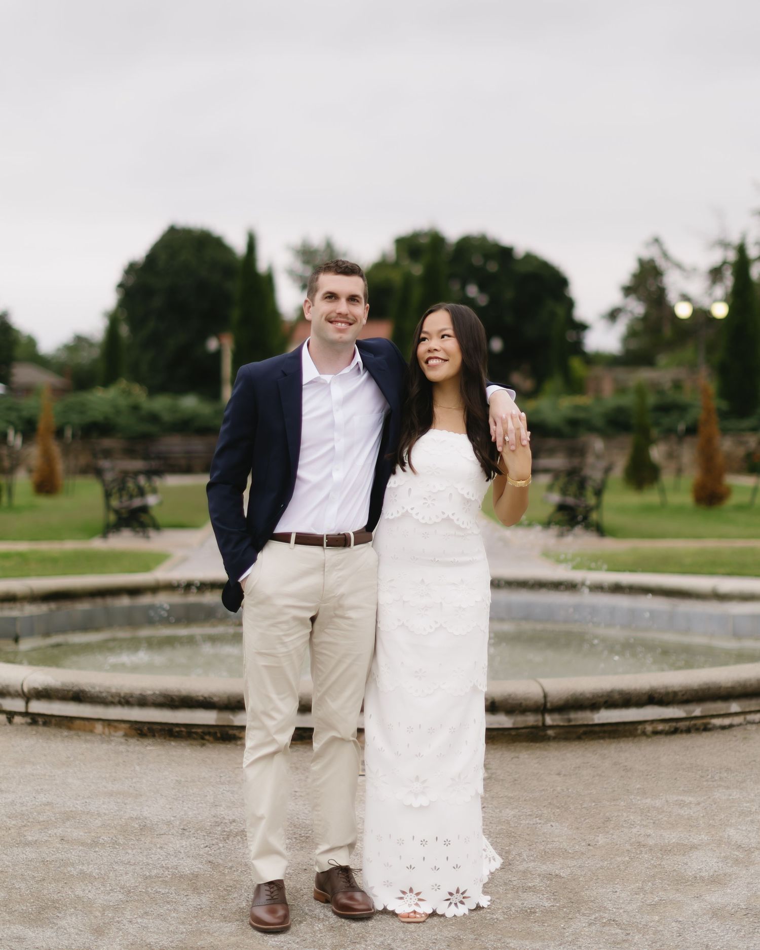 A couple in formal attire poses playfully in a garden setting with fountains visible in the background.