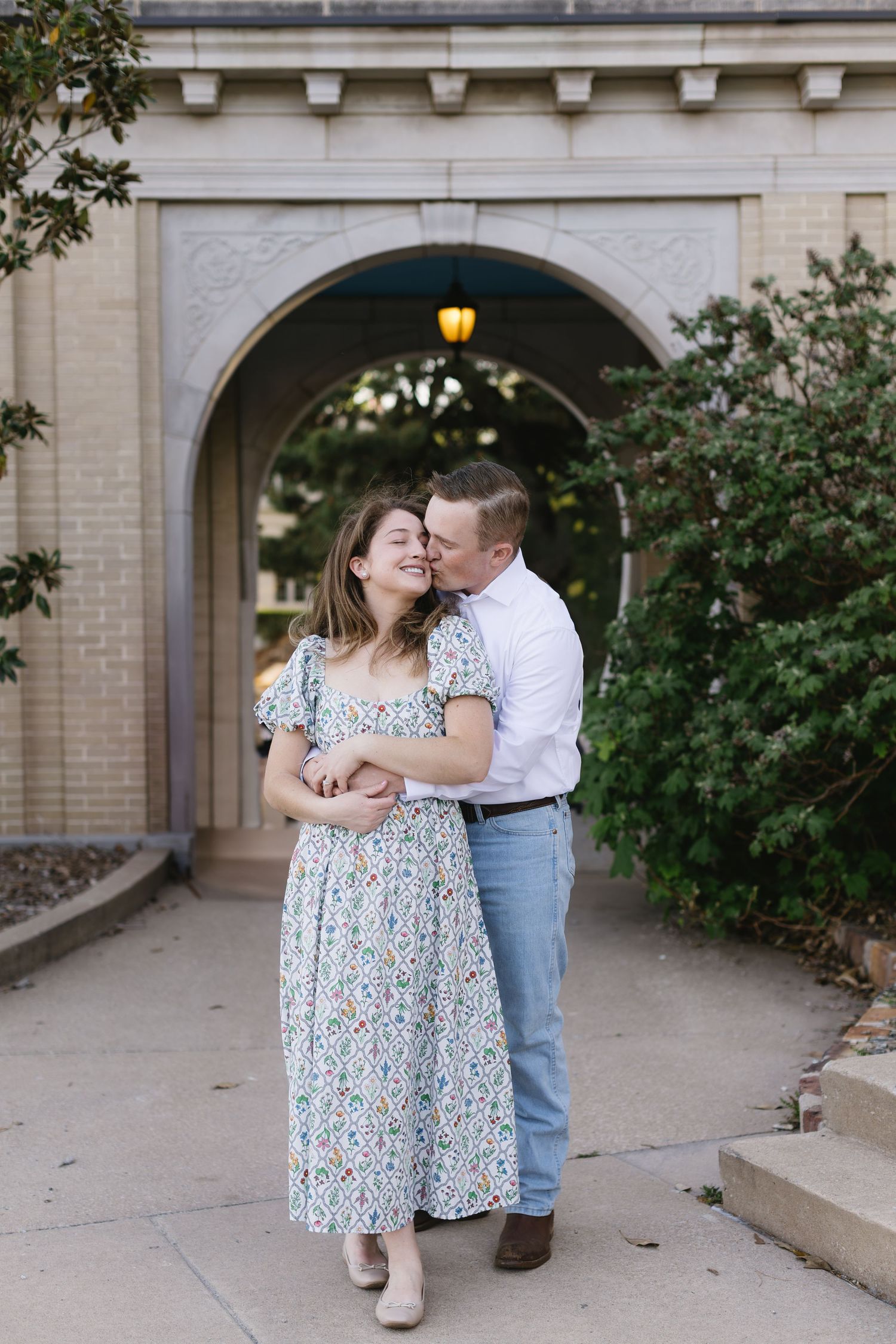 A romantic pair embraces under an archway with brick walls and greenery in the background.