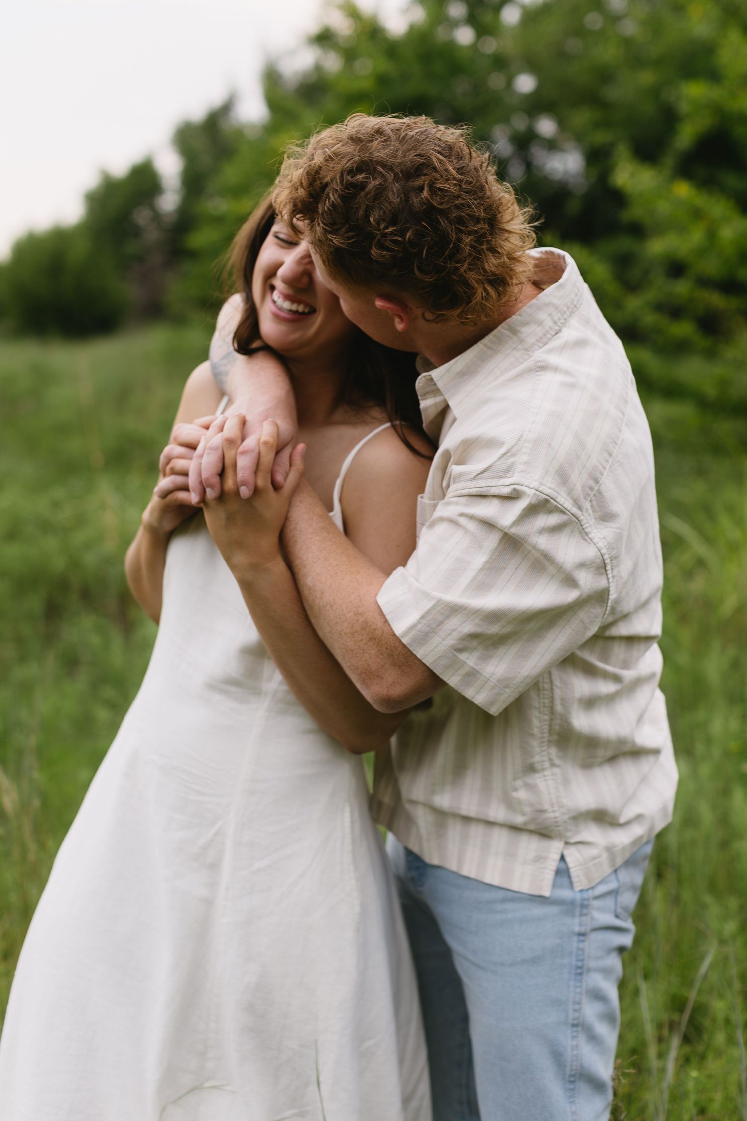 Romantic couple embraces in a lush green meadow during a photo session wearing light summer clothing.