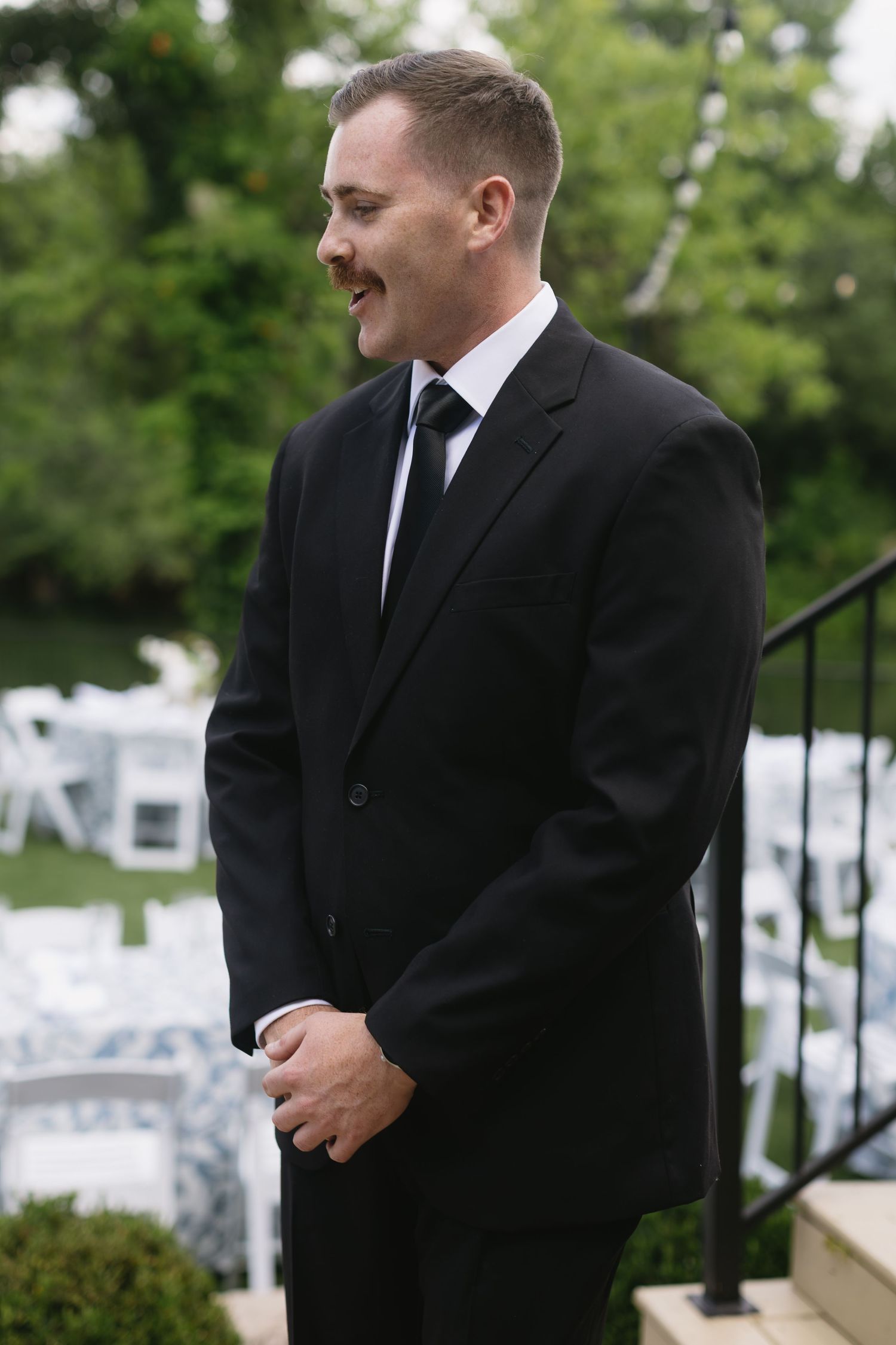 Groom in black suit standing outdoors with white chairs in background.