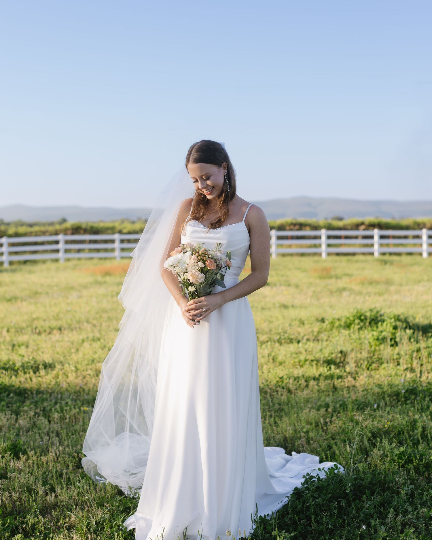 A bride in a white dress poses in a sunny meadow with a white fence and mountain views.