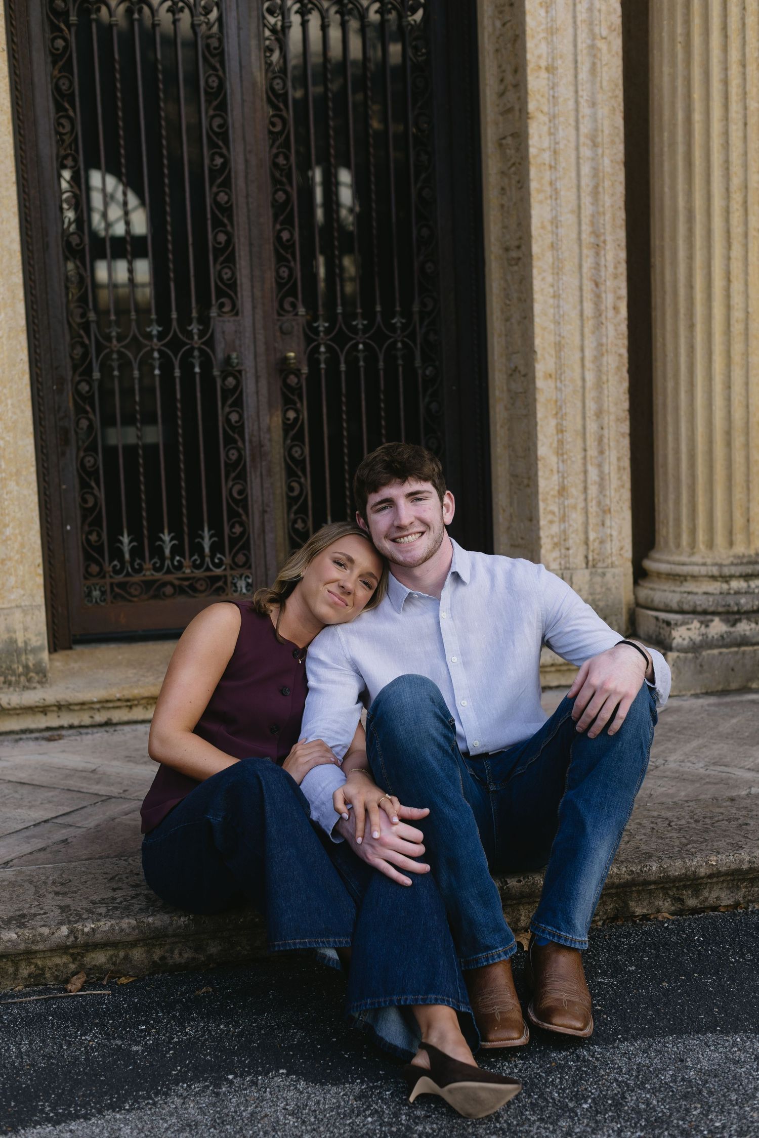 A casual couple sits together on stone steps by ornate iron gates, sharing a relaxed moment.