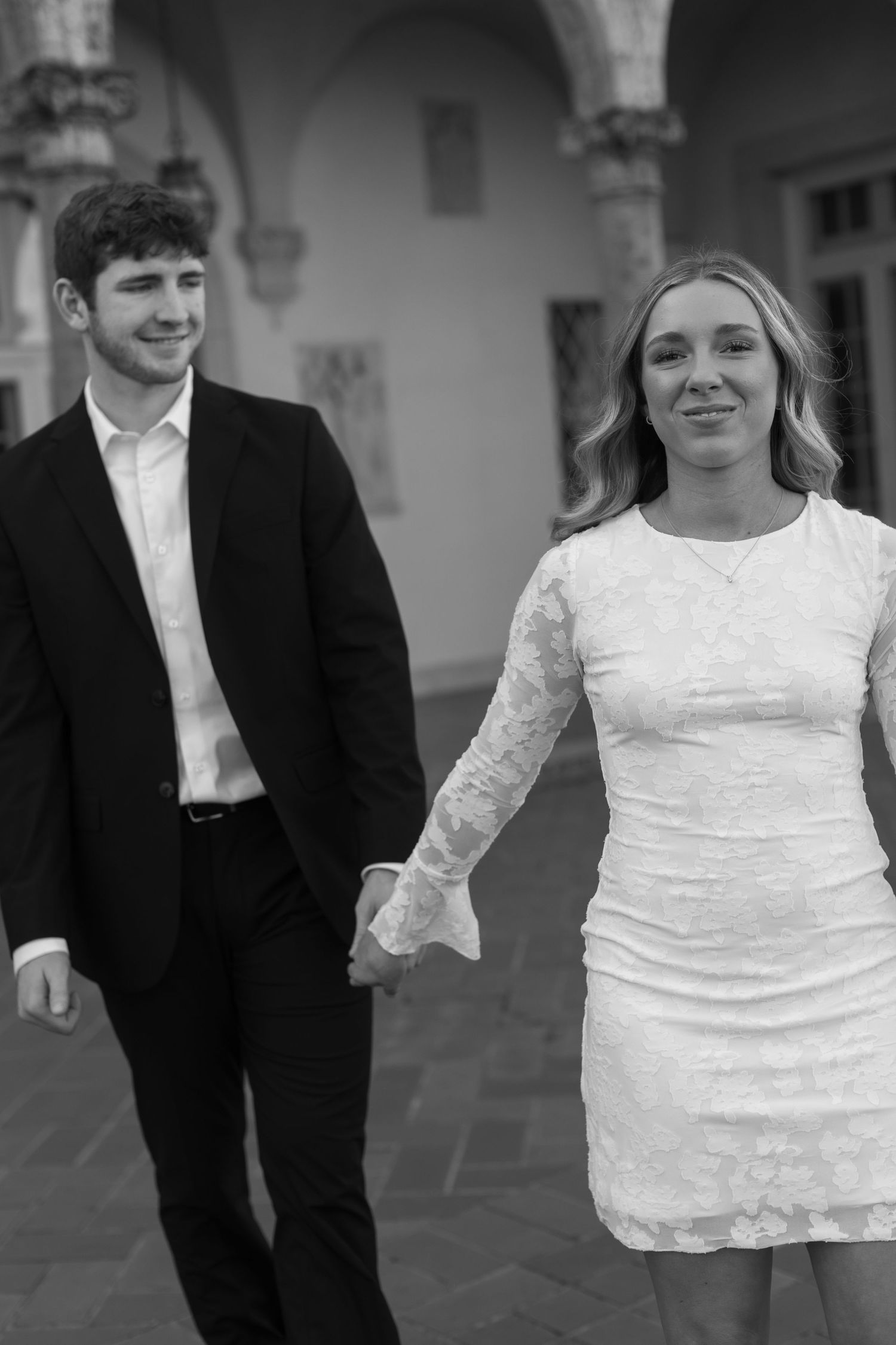 A couple in black and white formal wear holds hands while walking through an elegant courtyard.