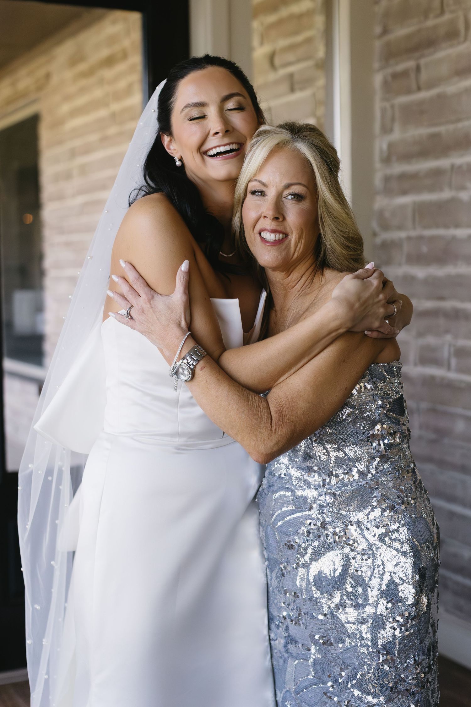 Joyful embrace between bride in white gown and woman in silver sequined dress.