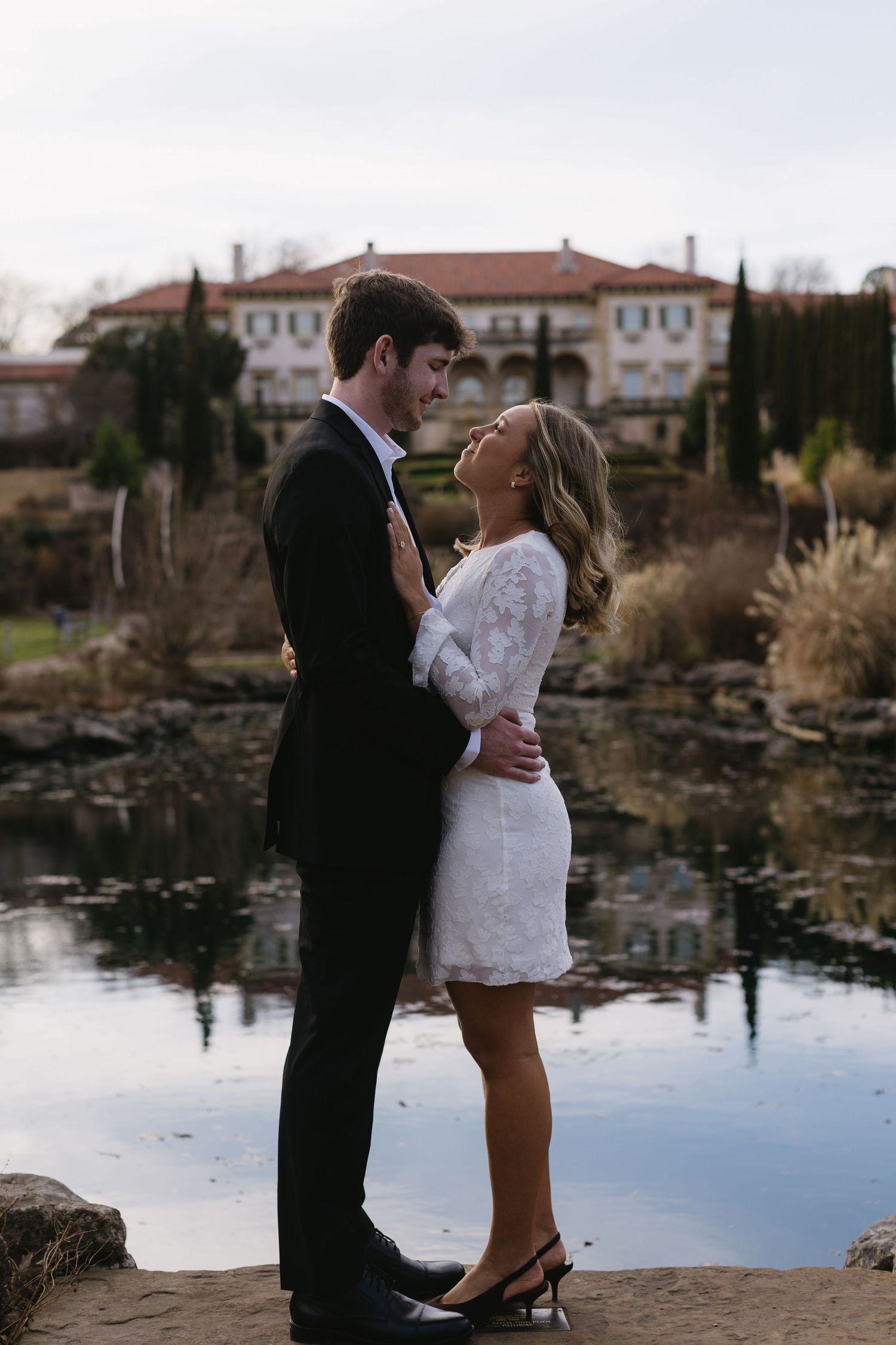 A couple poses romantically by a reflective pond with buildings visible in the background.