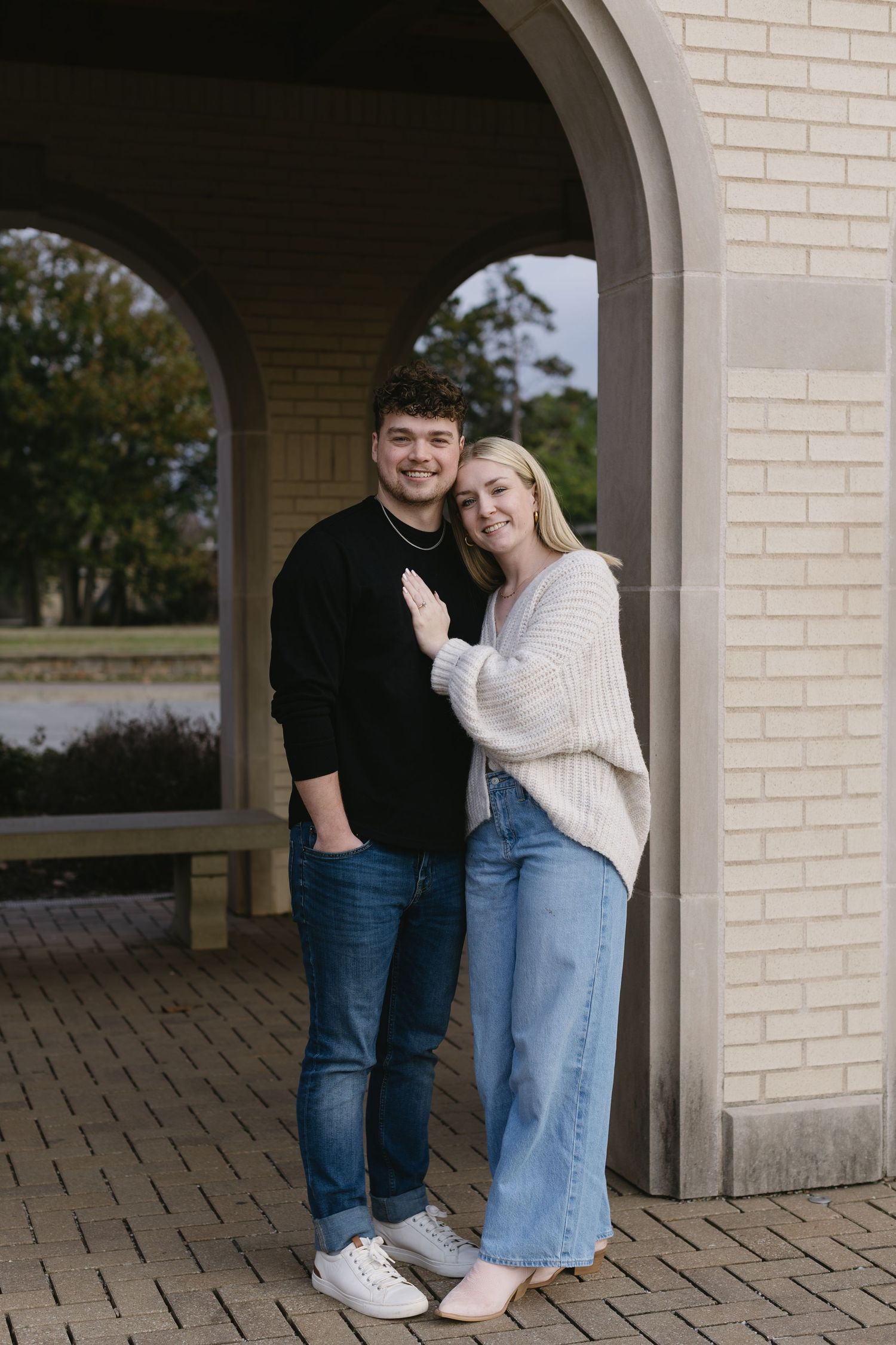 A couple poses together under brick archways wearing casual outfits of jeans and sweaters.