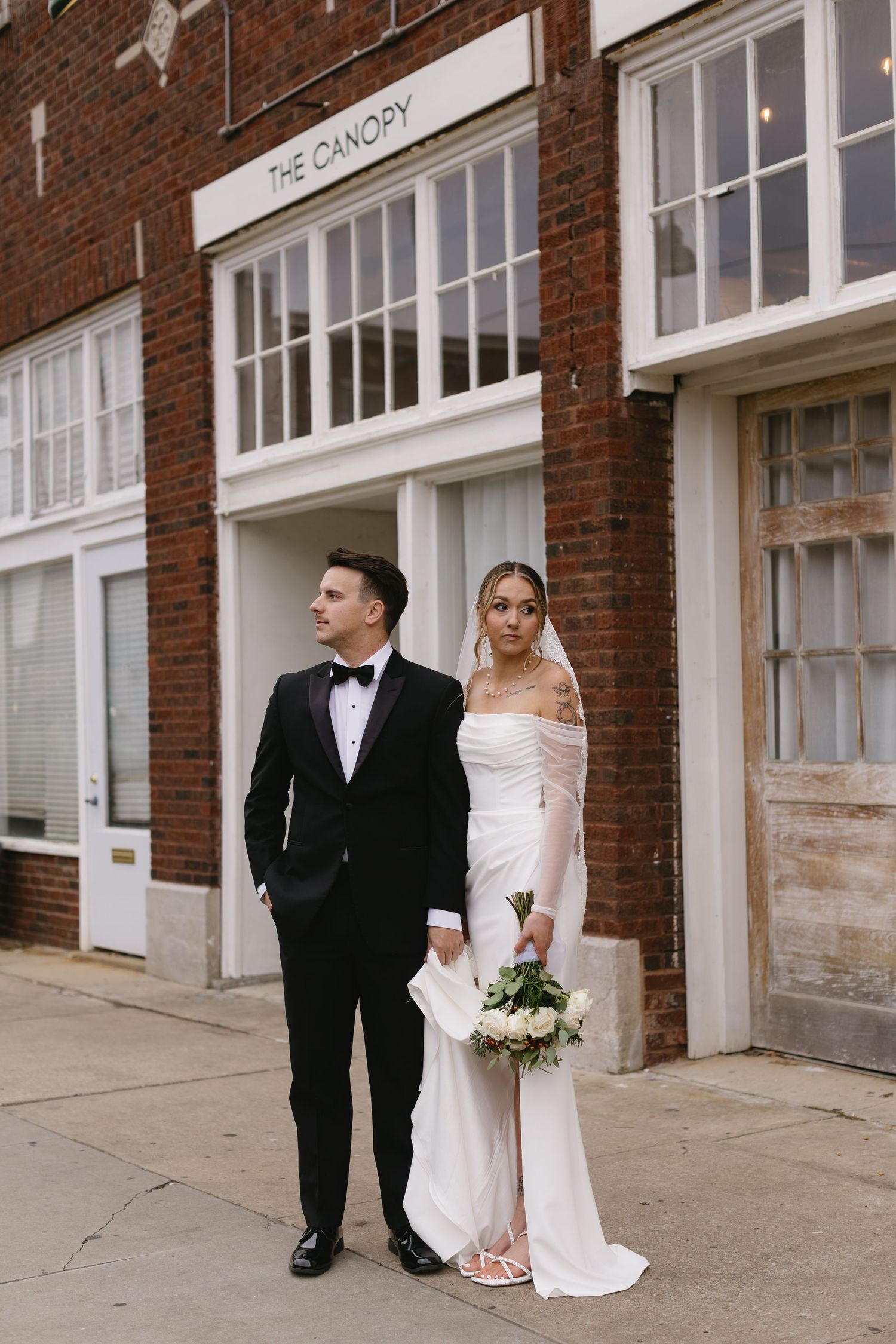 Elegant couple poses outside a brick building on their wedding day in classic formal attire.