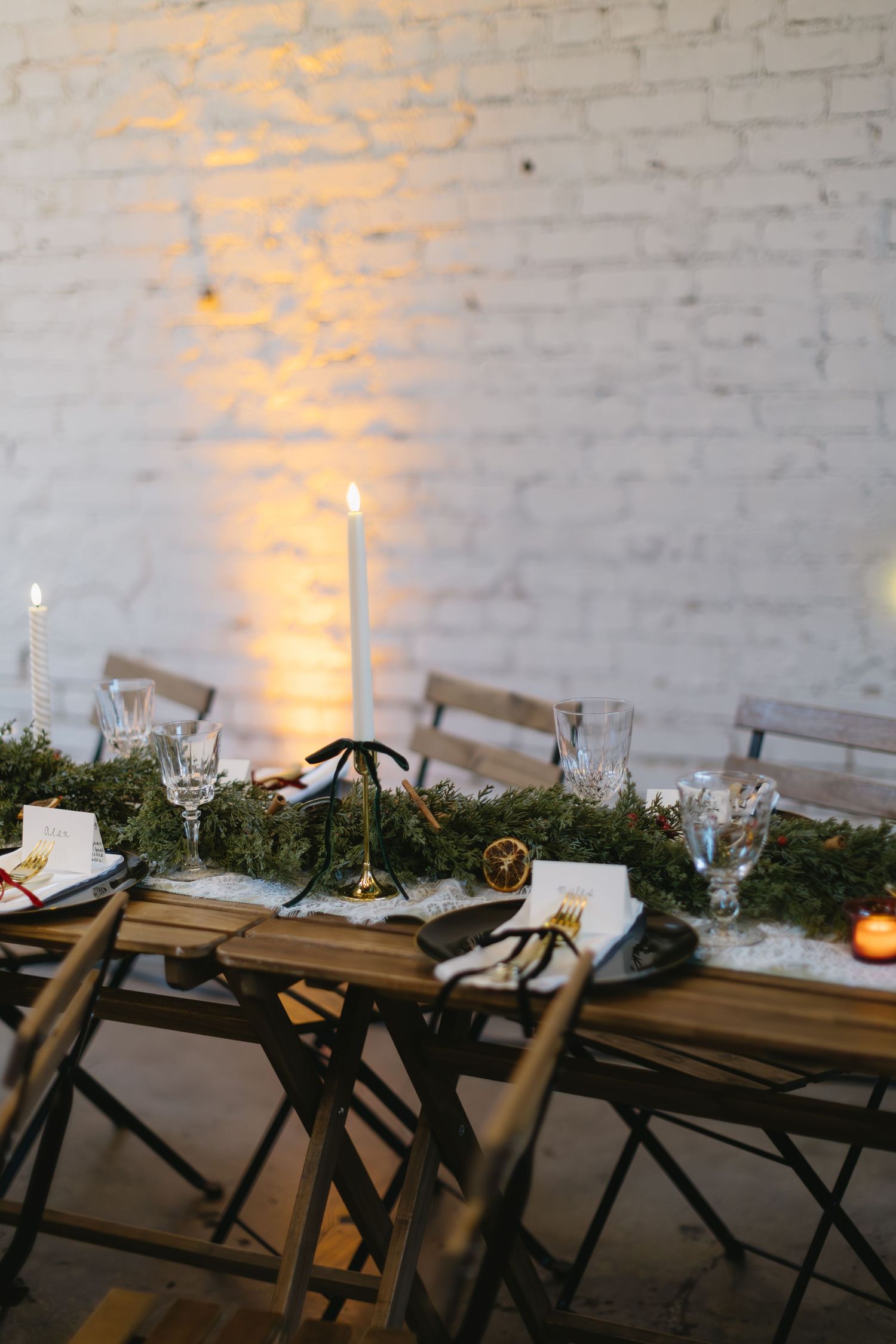 Long wooden table set for dinner with greenery, candles and place settings against white brick wall.