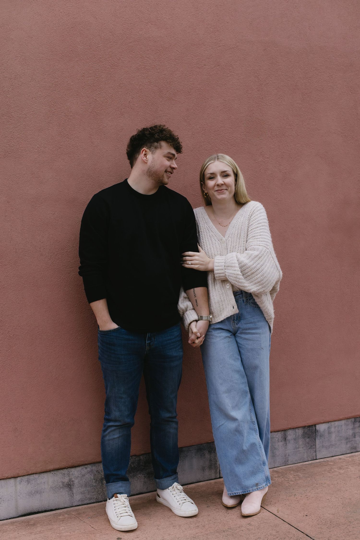 A couple in casual outfits leans against a terracotta colored wall during a photoshoot.