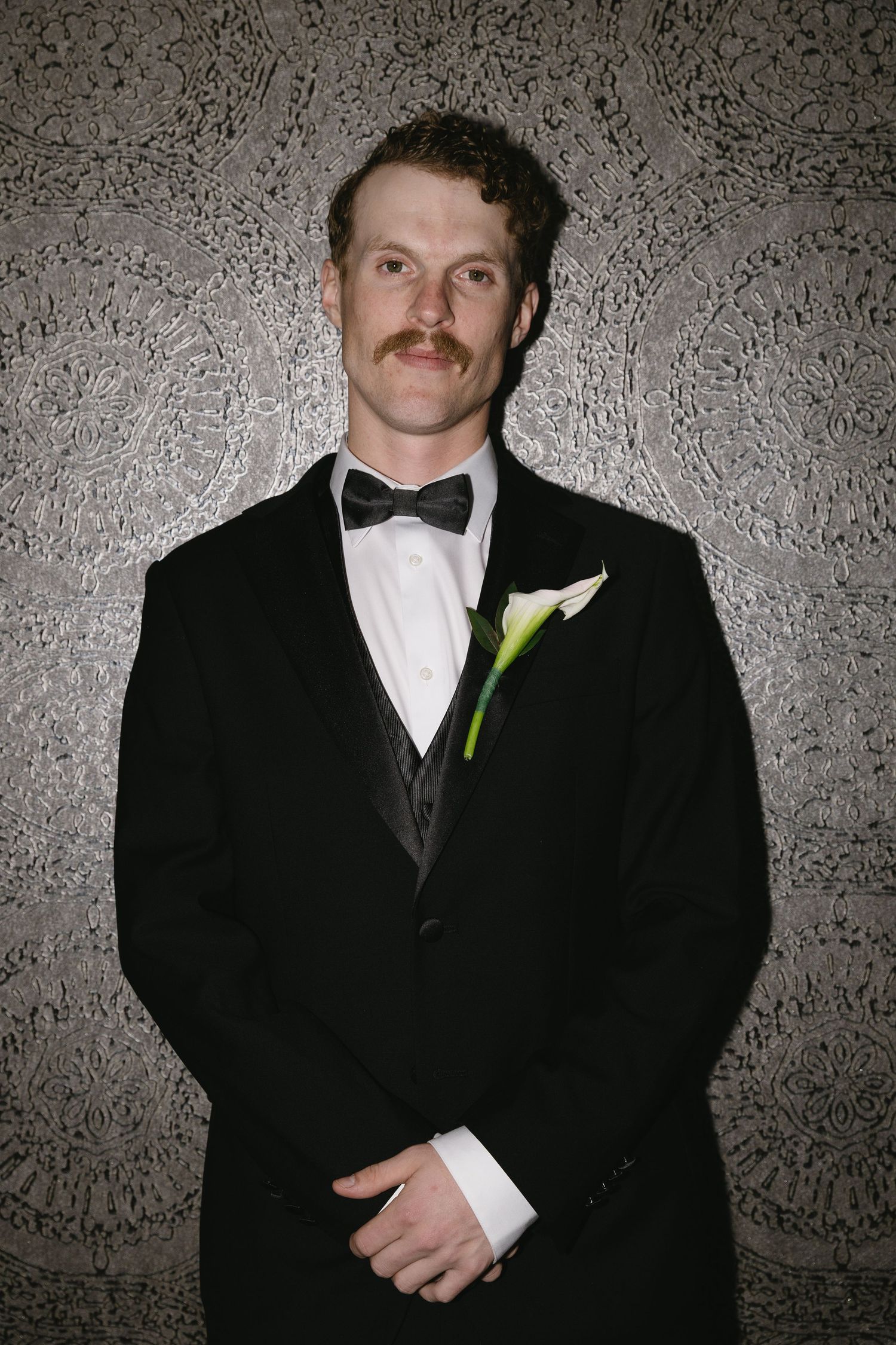 Formal portrait of a groom wearing a black tuxedo and white bow tie with a mustache against a textured wall.