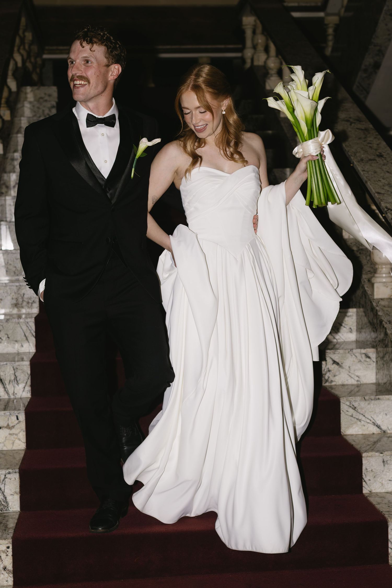 Wedding couple descending marble stairs with elegant white dress and tuxedo.