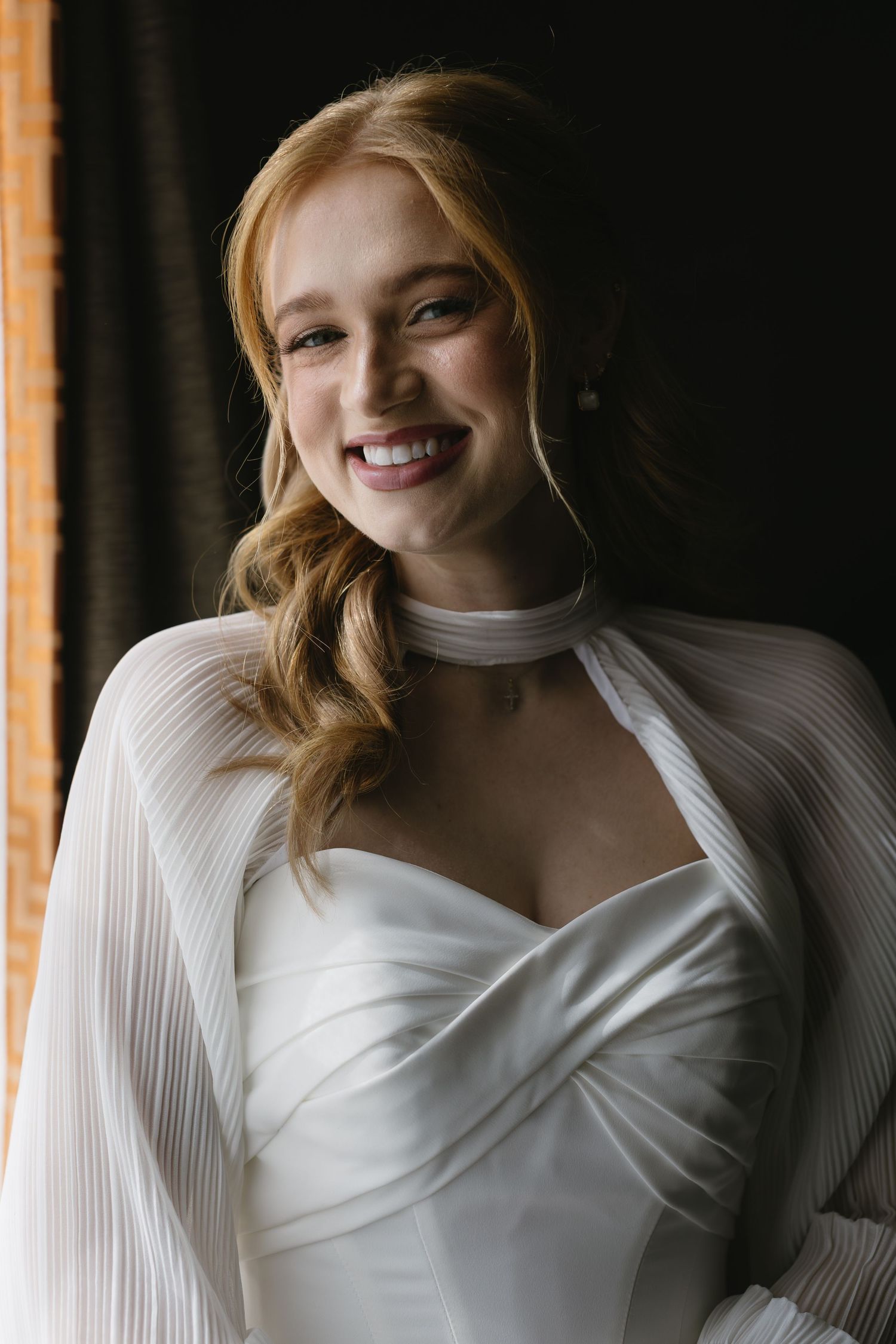 Bride wearing an elegant white pleated dress poses by window light with joyful expressions.