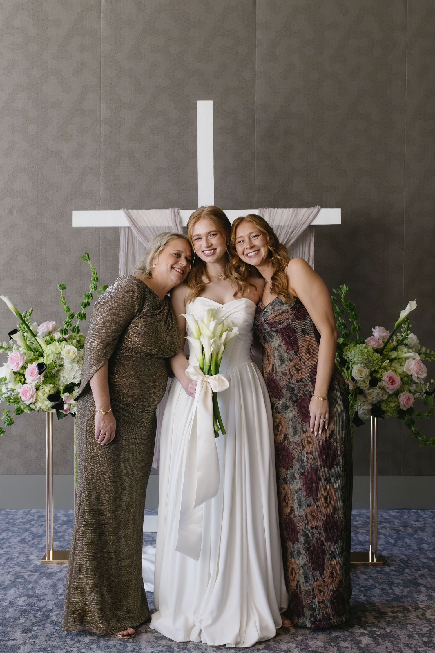 Wedding party poses against grey wall with floral arrangements and white cross backdrop.