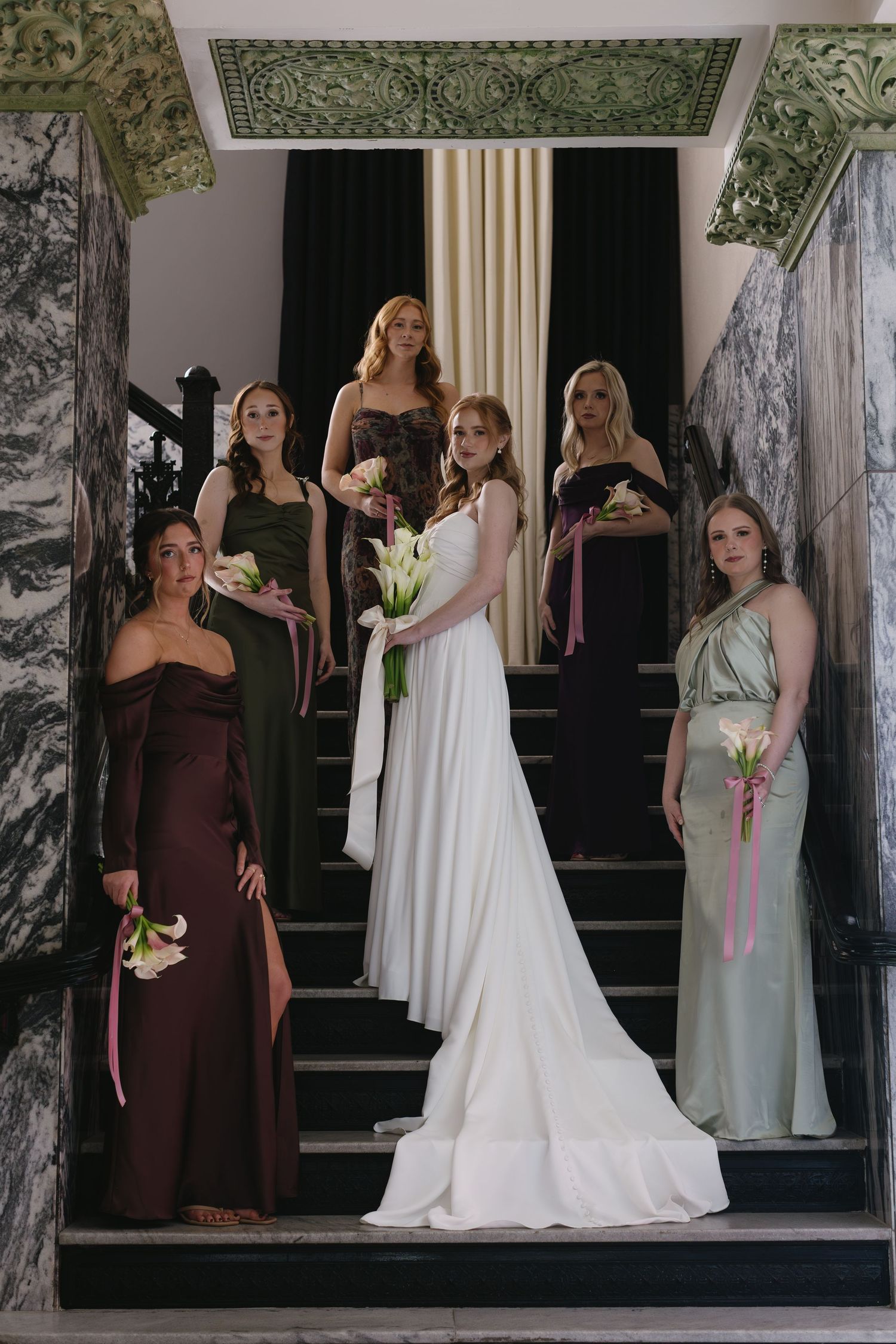 Wedding party members in brown and mint green dresses pose with bride in elegant white gown on stairs.