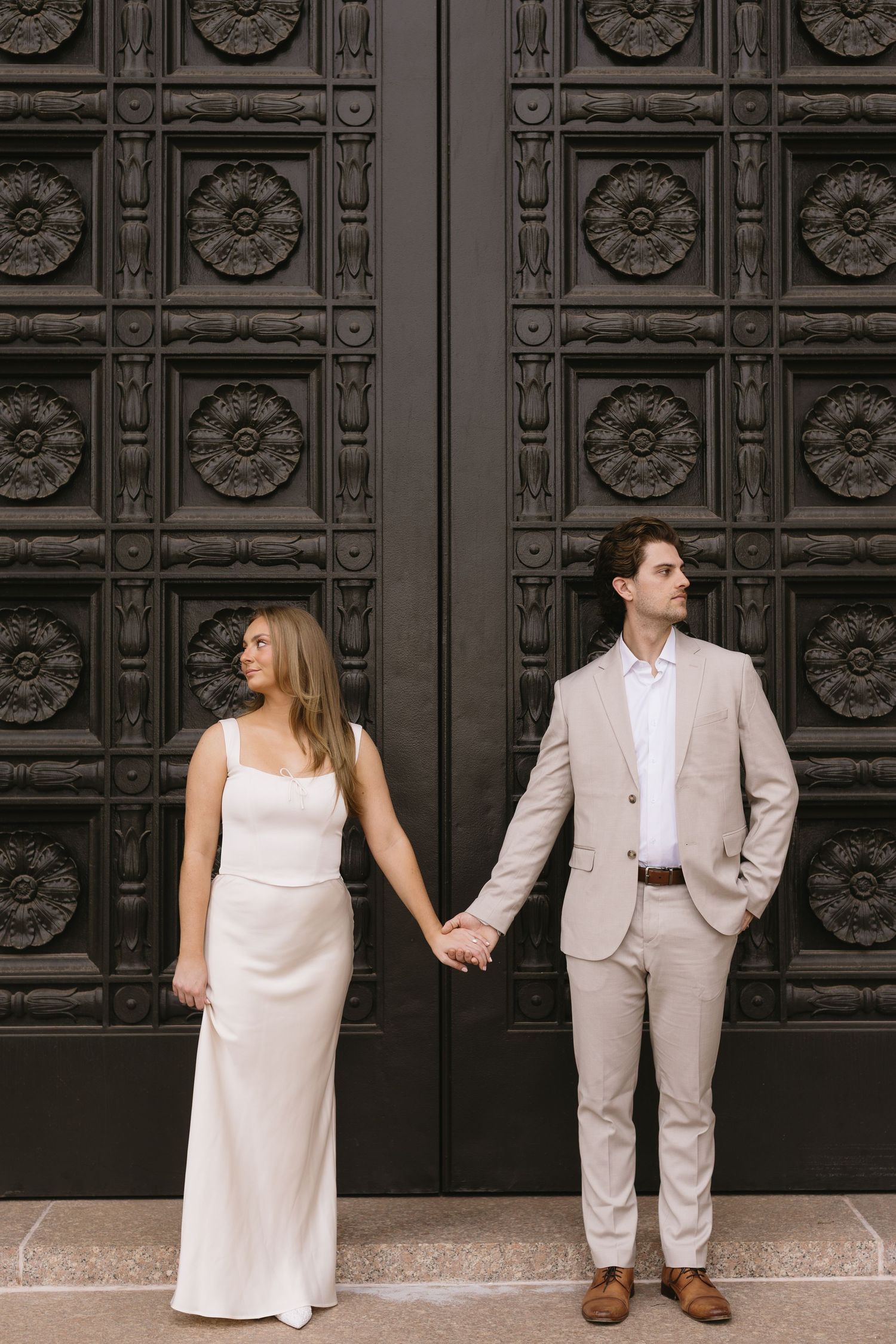 A couple in white dress and beige suit poses against ornate black doors of a historic building.