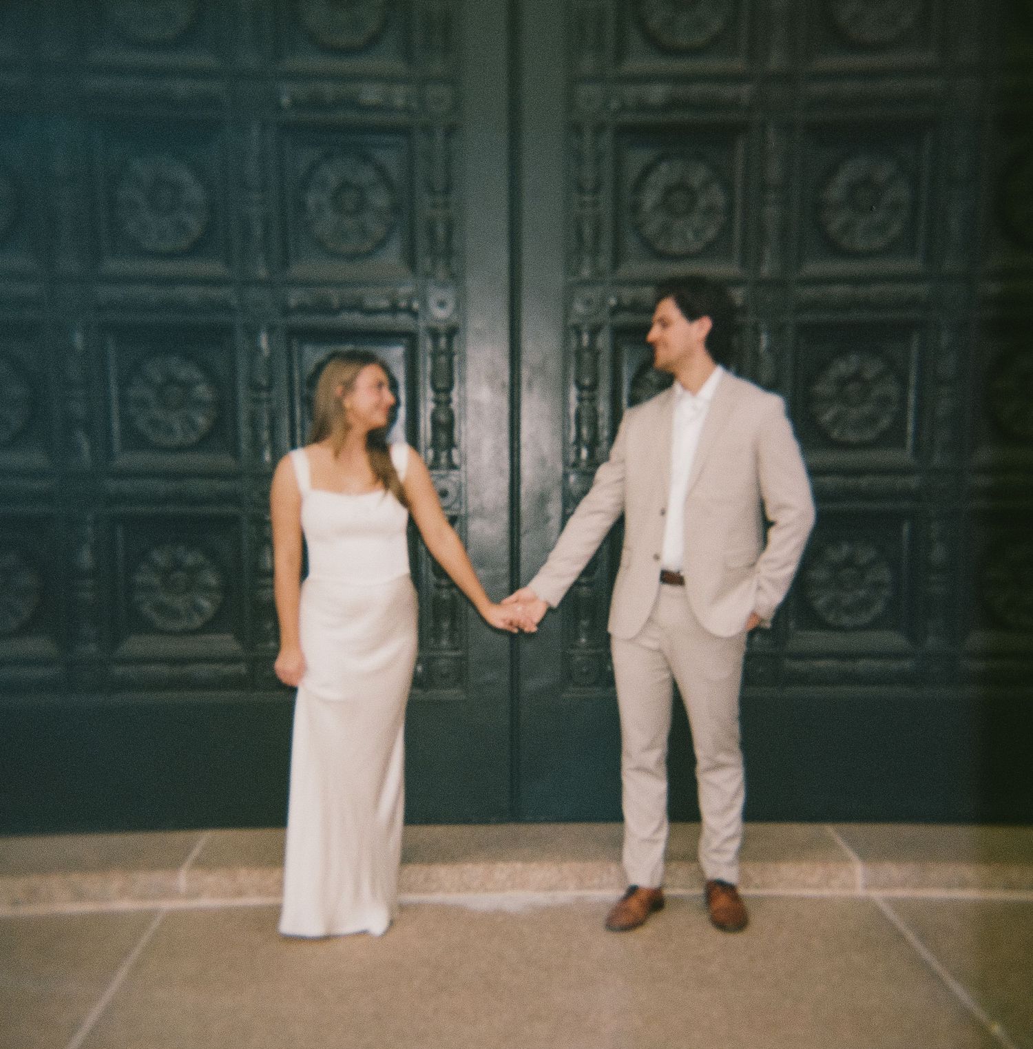 A couple in formal attire poses in front of ornate metal doors.