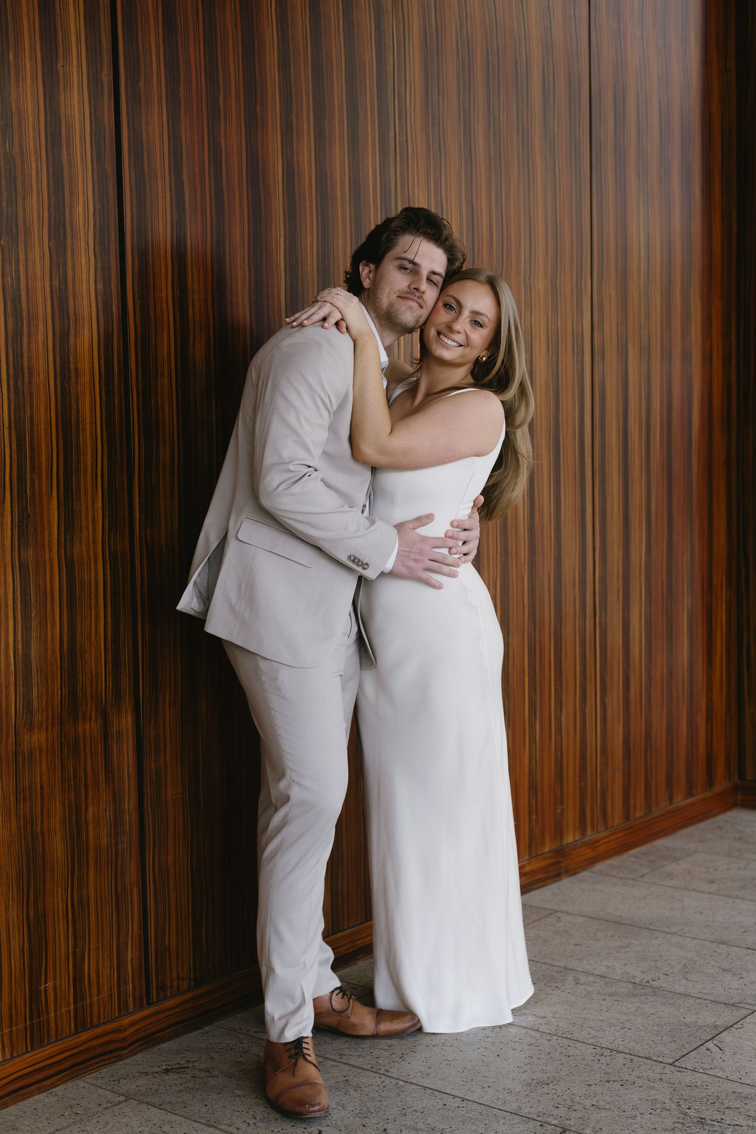 Couple in white and beige formal wear embraces against wood-paneled wall.