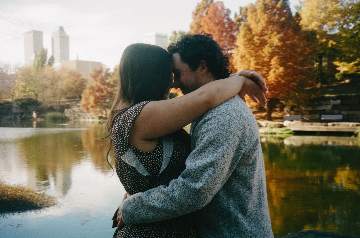A couple embraces near a tranquil lake surrounded by autumn trees with golden foliage in a romantic outdoor setting.