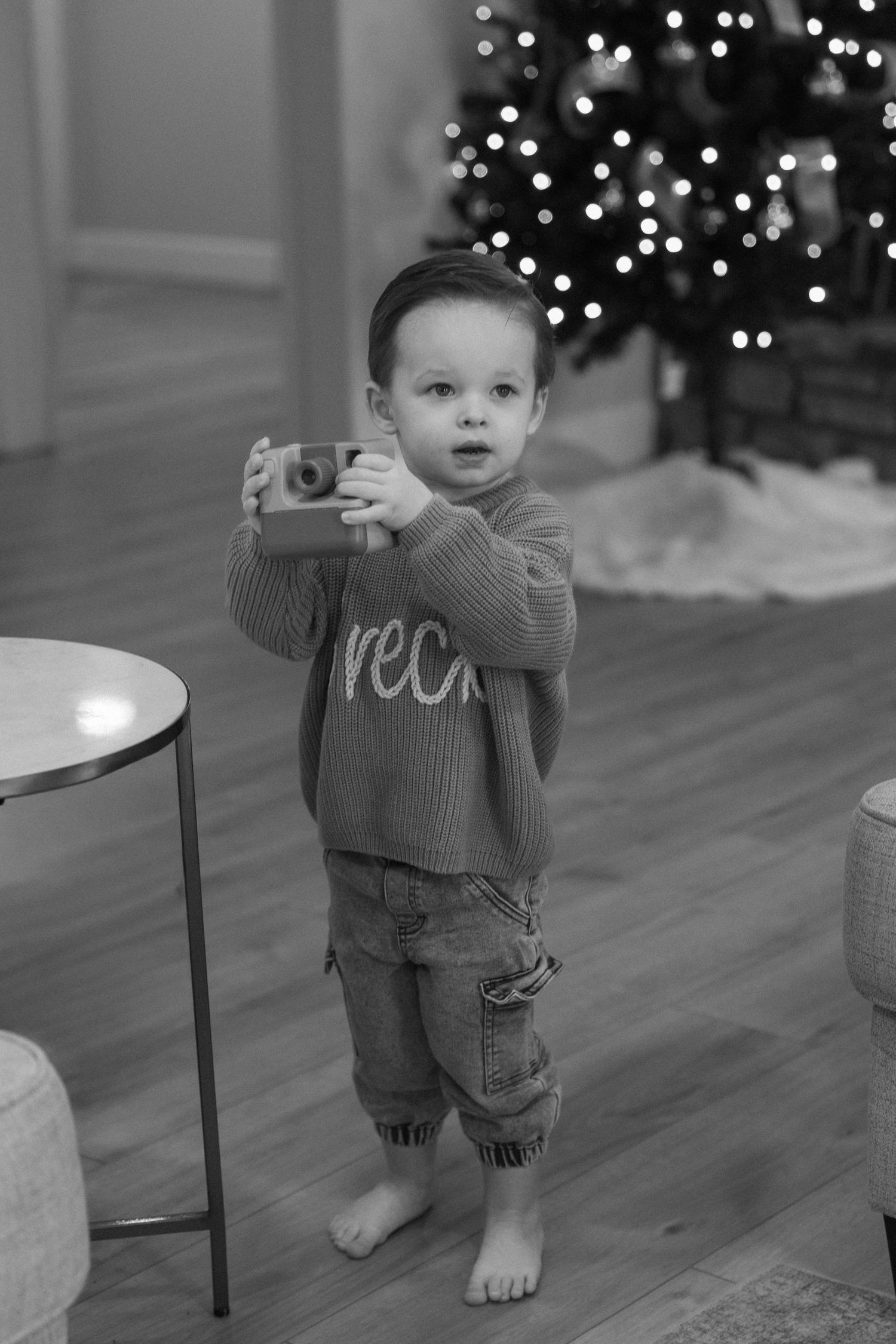 Small child in grey sweater standing and holding onto chair near Christmas tree lights.