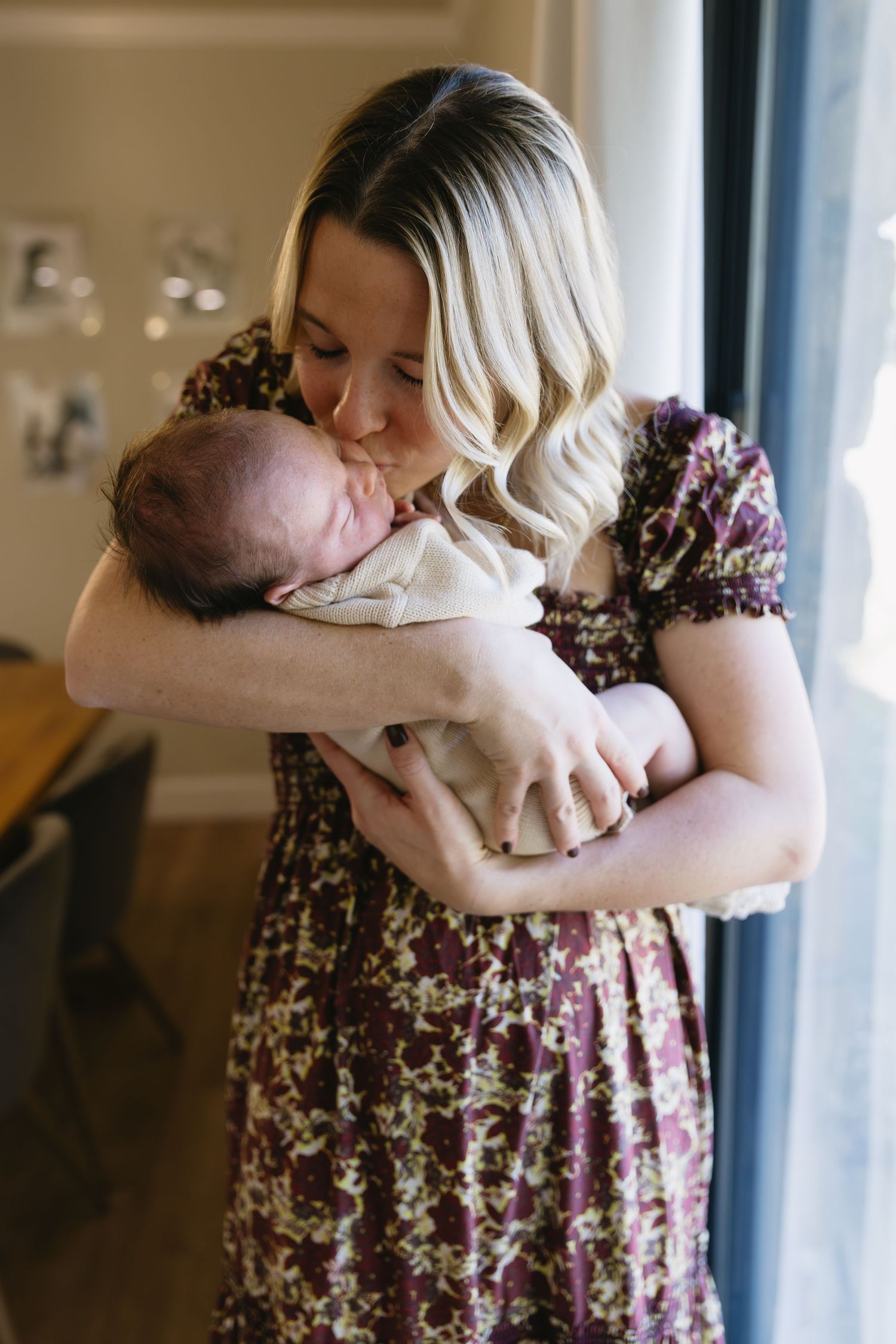 A tender moment showing a floral dress-clad figure holding a small infant near a window.