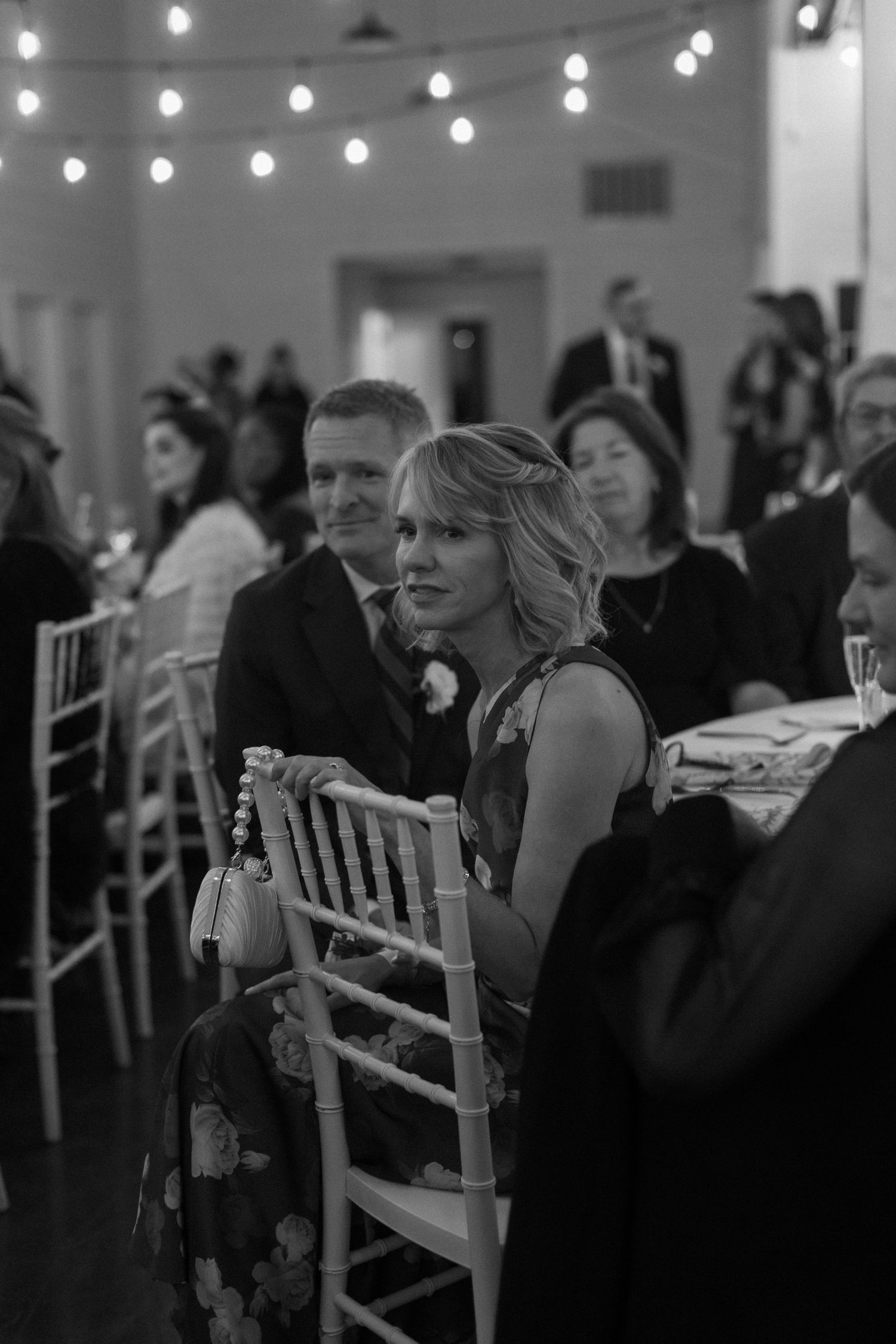 Wedding reception guests seated at tables with string lights overhead in black and white.