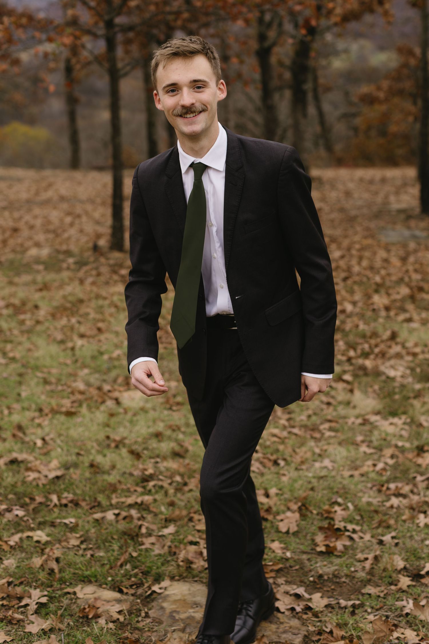 A person in a black suit and tie walks through a fall landscape covered in autumn leaves.