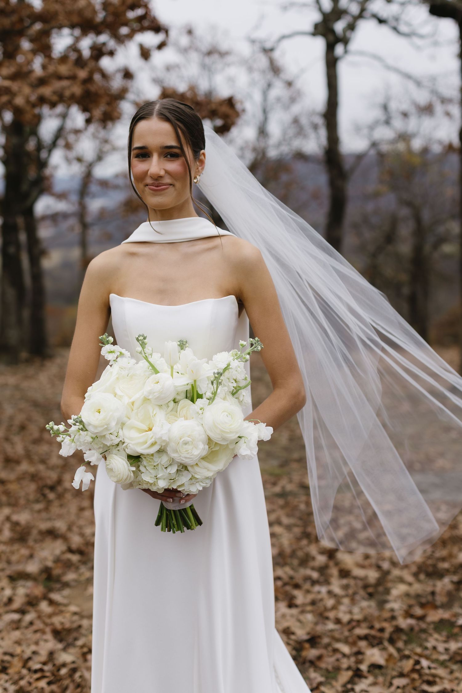 Elegant bridal portrait with white bouquet and flowing veil in autumn setting.