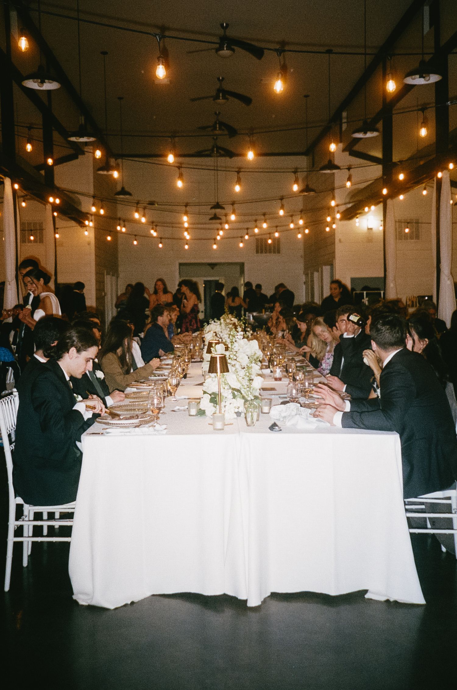 Wedding reception venue with long tables, white linens, and string lights creating an elegant ambiance.