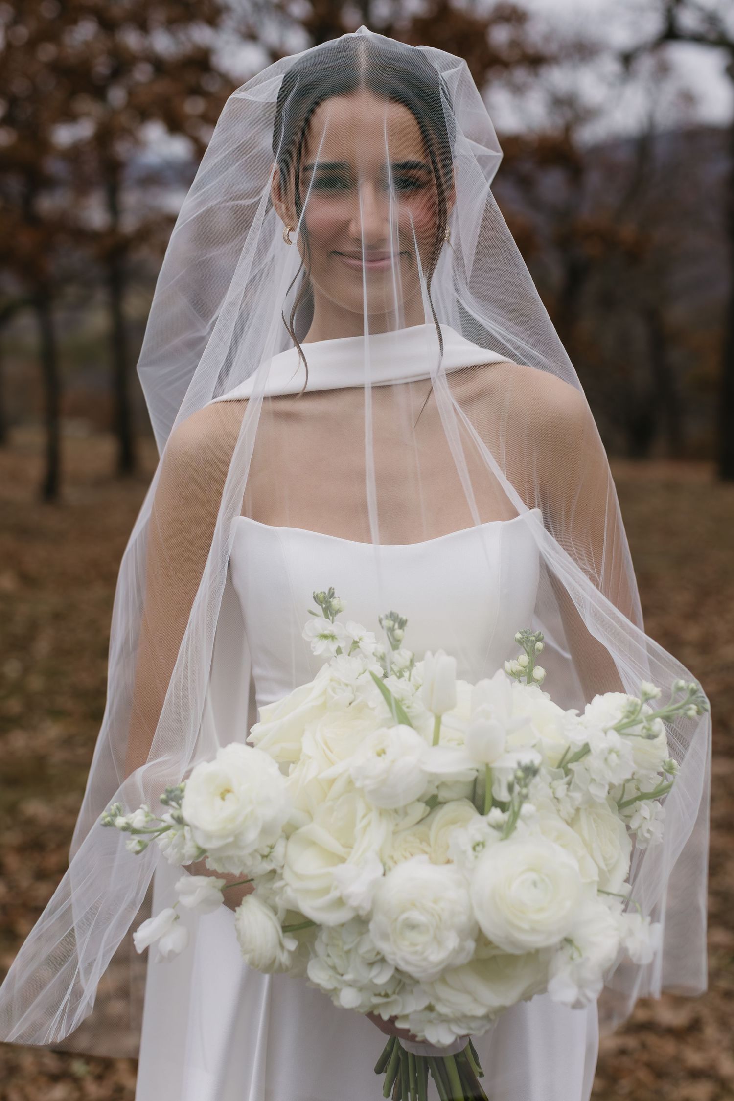 Bride in white dress holds stunning white floral bouquet during fall outdoor photoshoot.