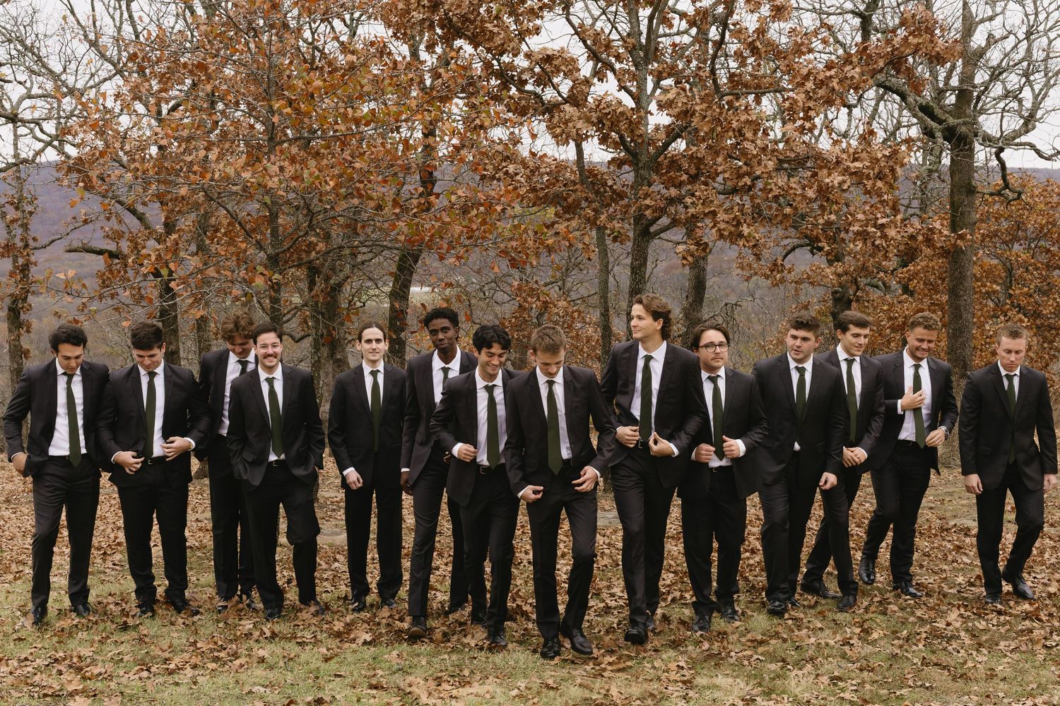 Groomsmen in matching black suits and ties pose together against an autumn backdrop with brown trees.