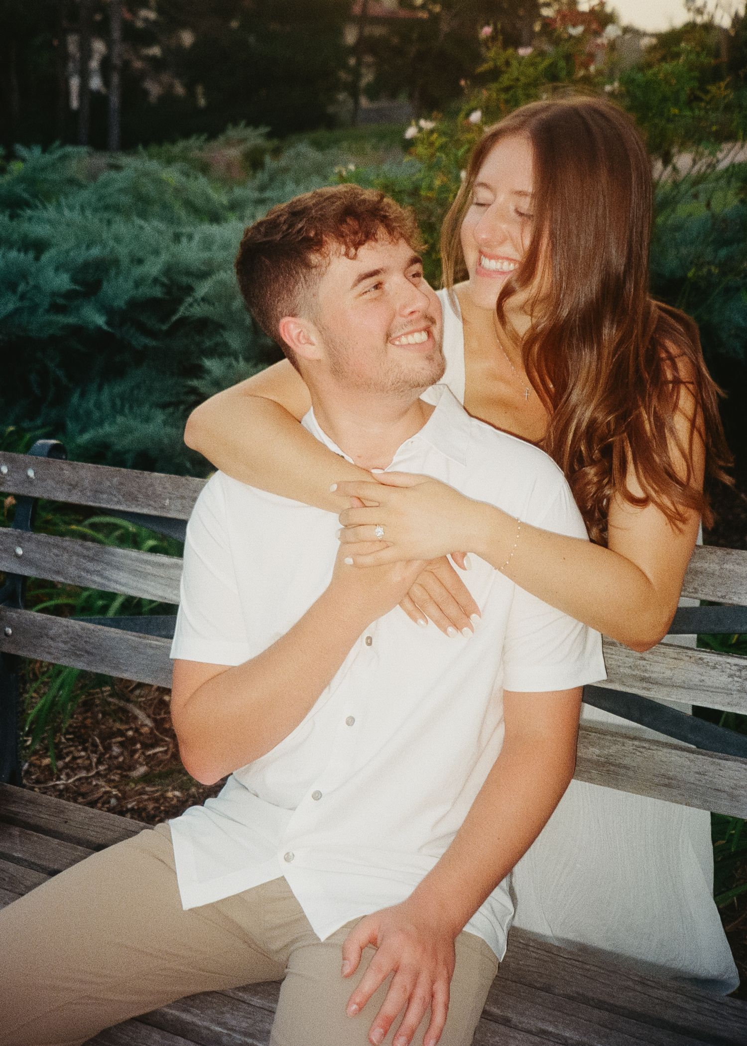 A joyful couple in white shirts share a candid laughing moment together on a wooden bench in a garden setting.