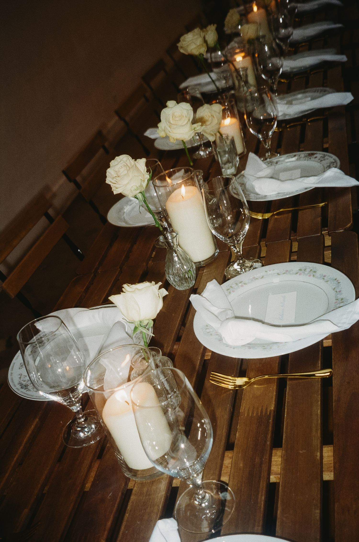 Elegant table setting with white roses, candles and crystal glassware on wooden slats.
