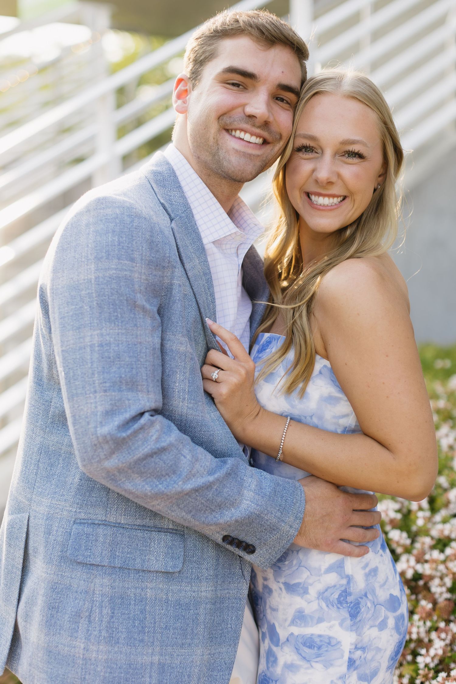 Romantic engagement photos capture a couple in light blue and white attire against a white wall with flowers.