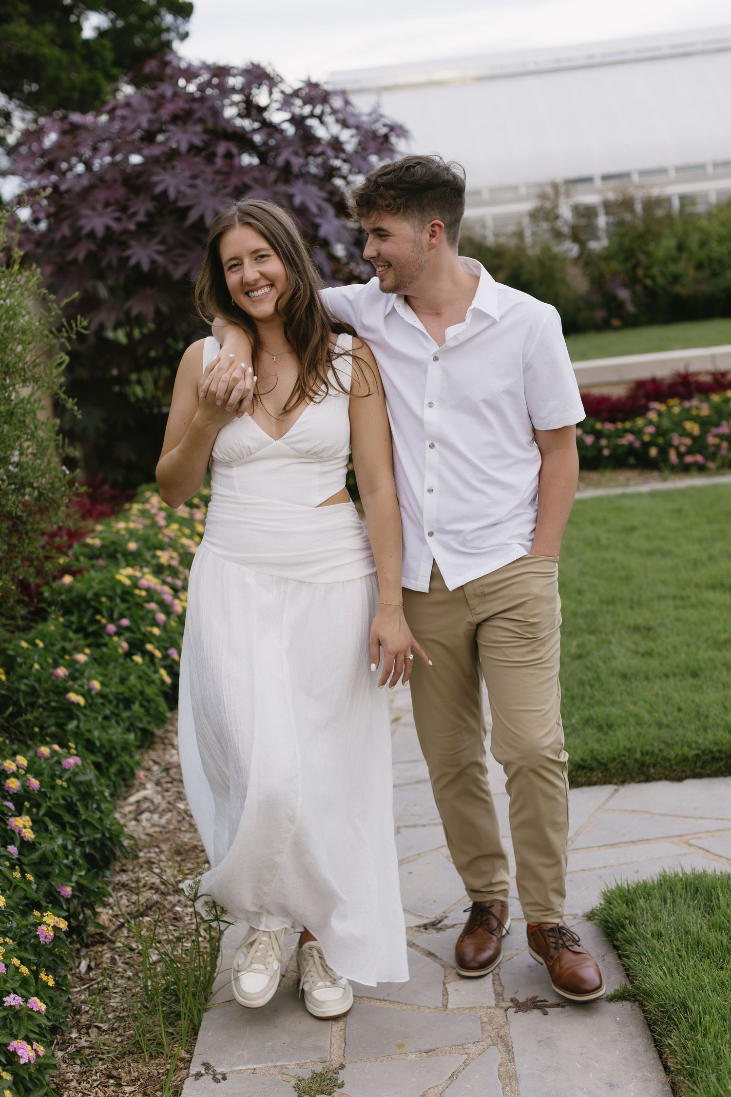 A couple walking together on a garden path, with her in a white dress and him in casual attire.