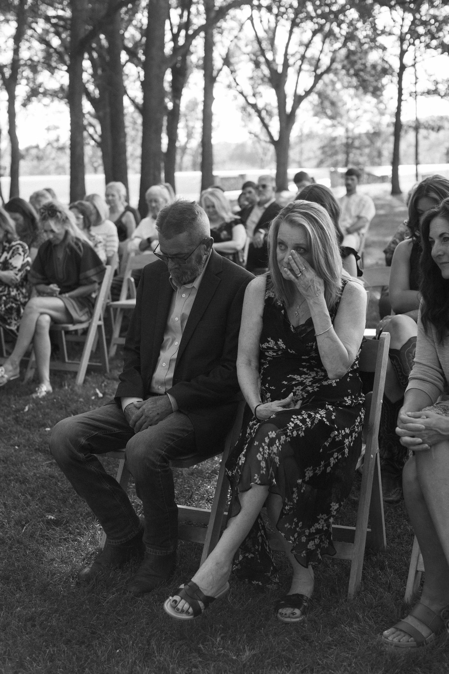 Wedding guests sit in rows of chairs outdoors among trees during an emotional moment at a ceremony.
