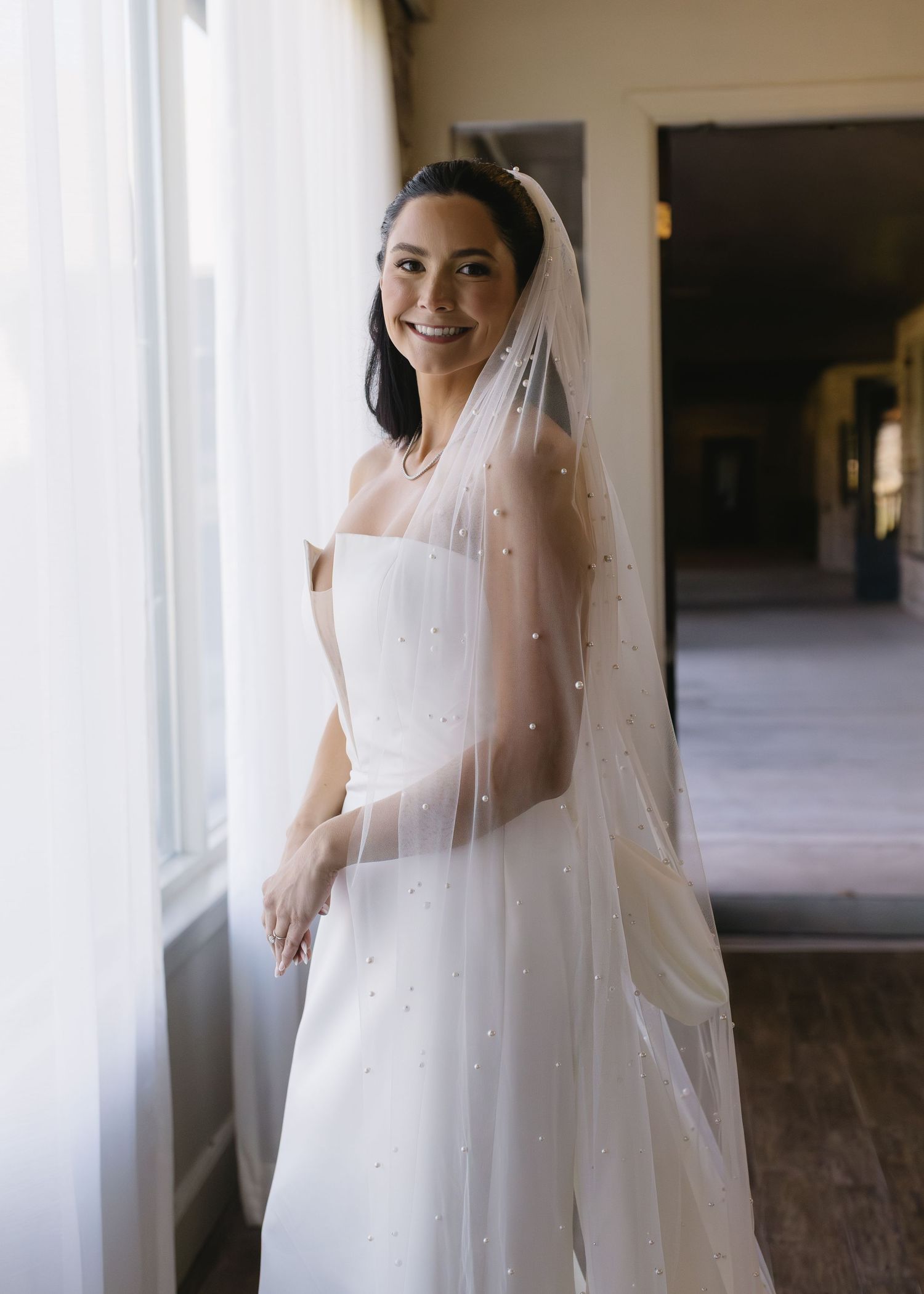 A bride in a flowing white dress with pearl details and veil stands in a sunlit hallway.