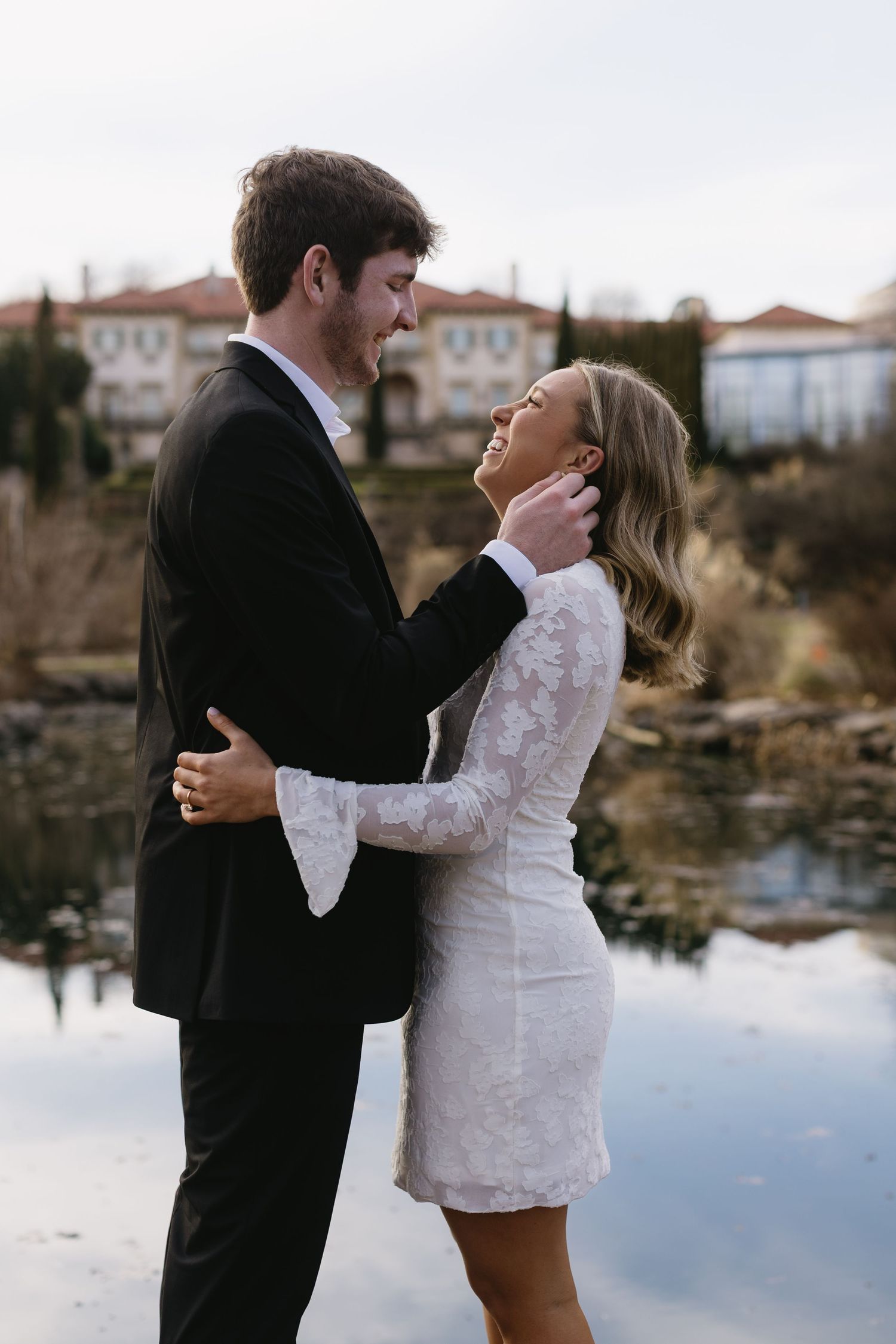 A couple shares an intimate moment by a reflective pond with buildings in the background.