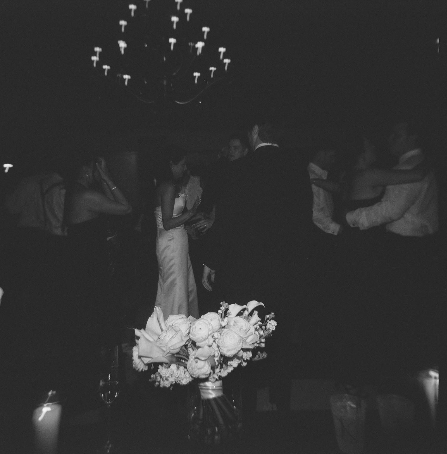 Black and white photo of couples dancing at an elegant wedding reception under a chandelier.