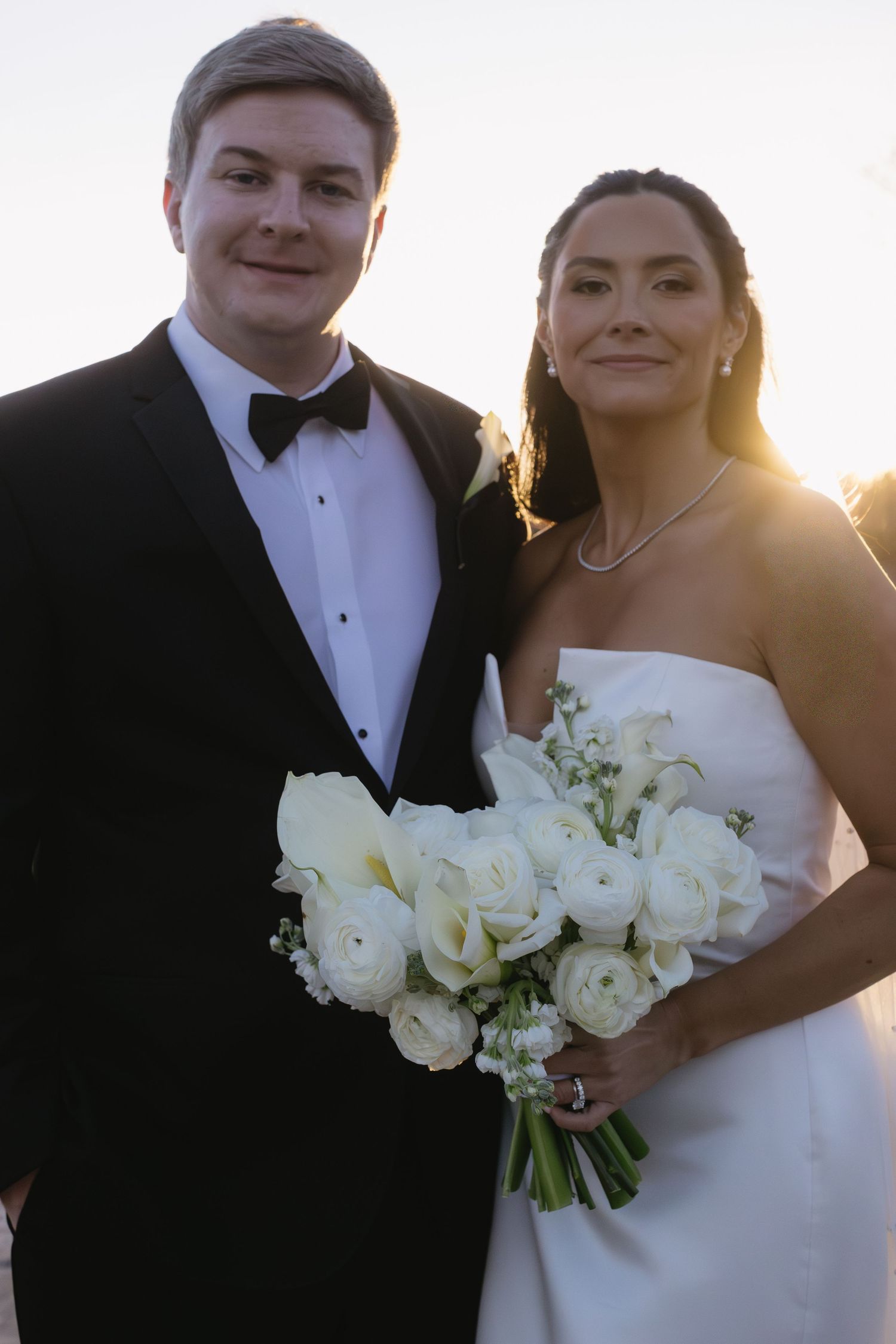 A couple in wedding attire posing with a white rose bouquet at sunset.