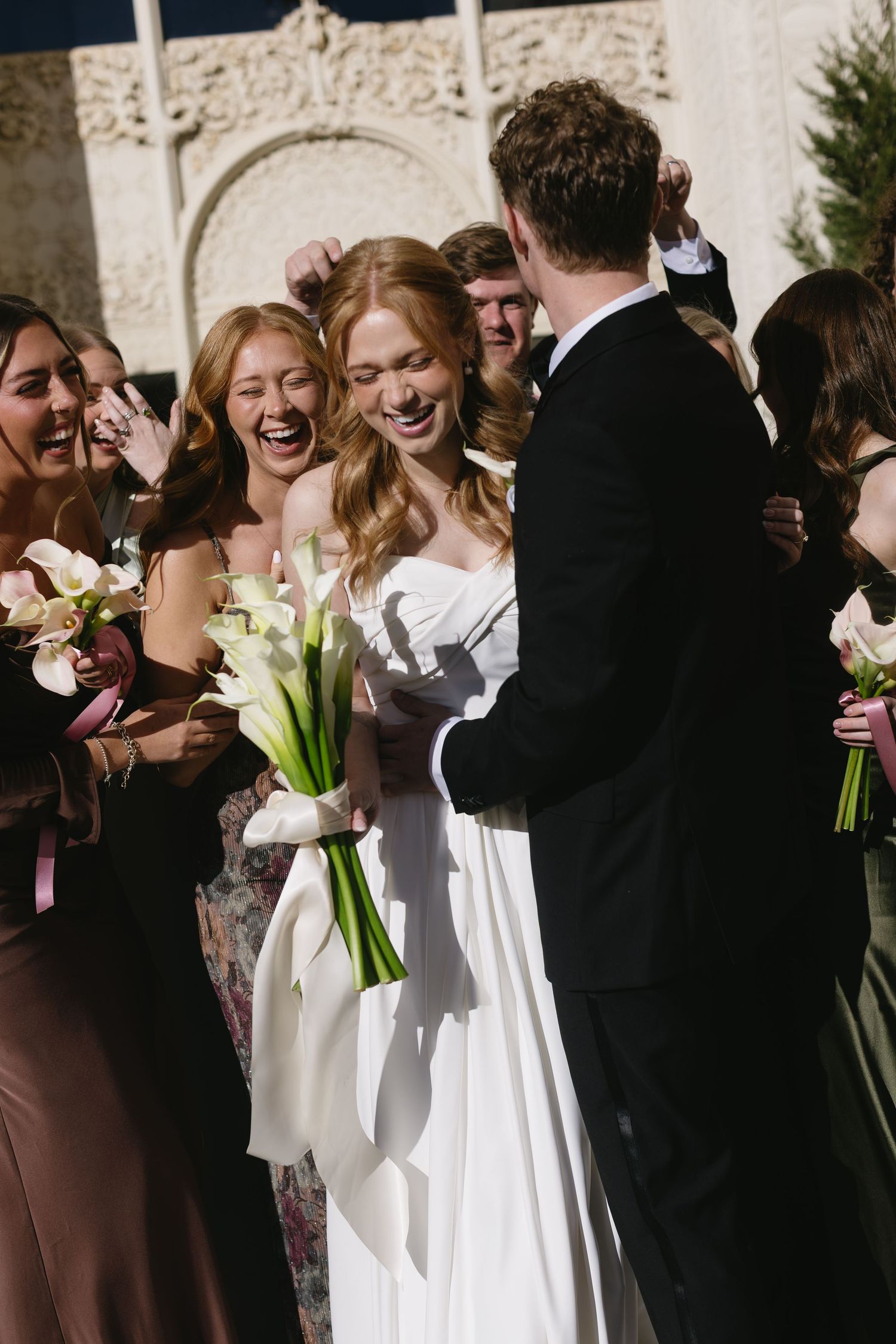 Wedding party gathered outside sharing joyful moments with bridesmaids holding white bouquets.