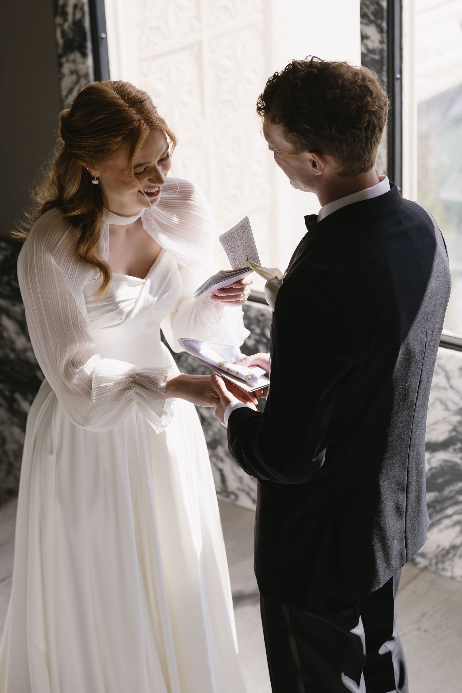 Wedding moment captured as couple exchanges notes in marble-walled setting.