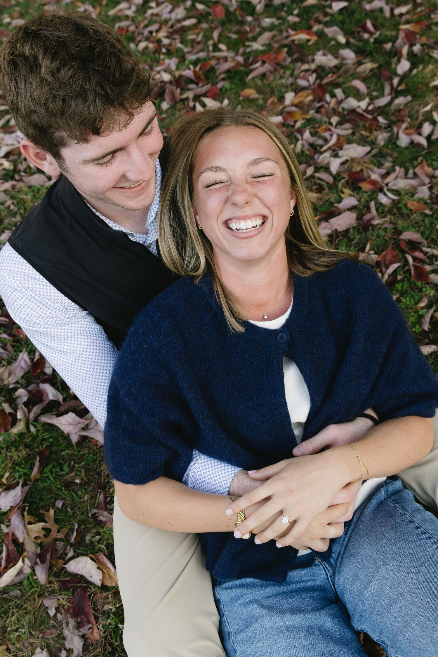 A couple sharing a candid moment of laughter while sitting on autumn leaves in a garden setting.