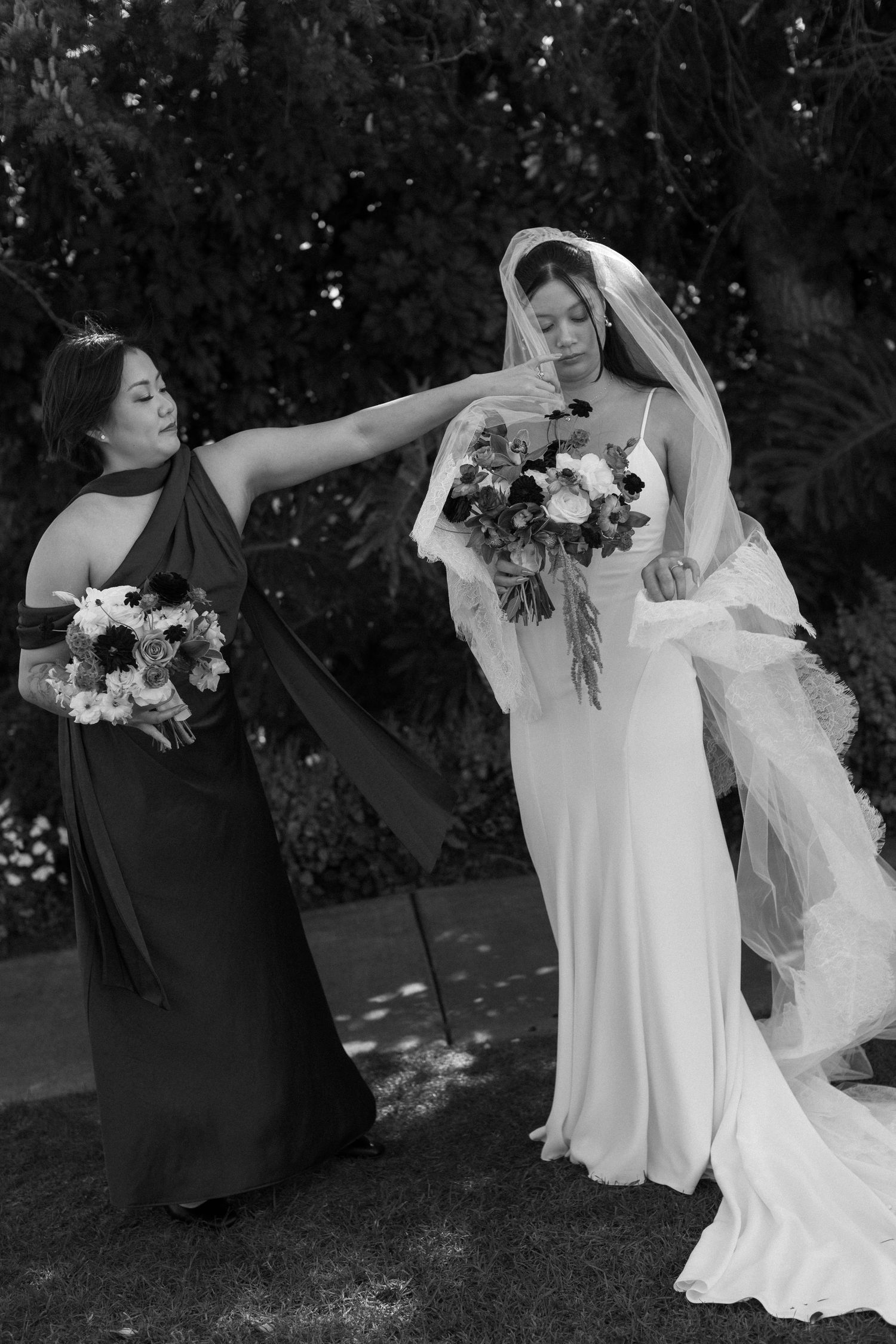 A bridesmaid in black adjusts the veil of a bride in a flowing white dress holding a floral bouquet outdoors.