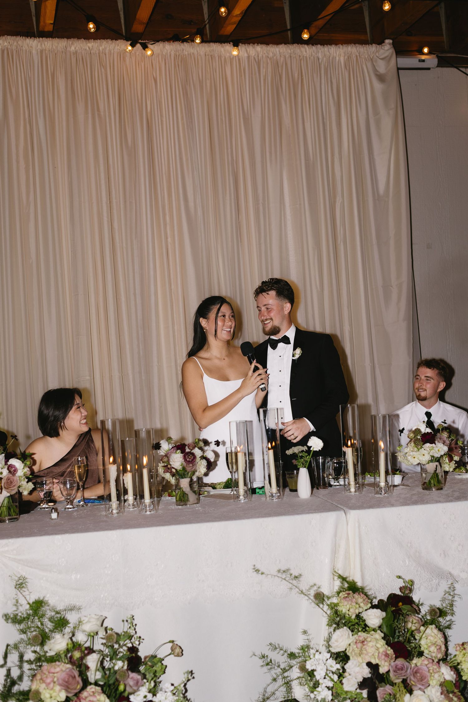 Wedding reception head table decorated with floral arrangements and candles as newlyweds celebrate.