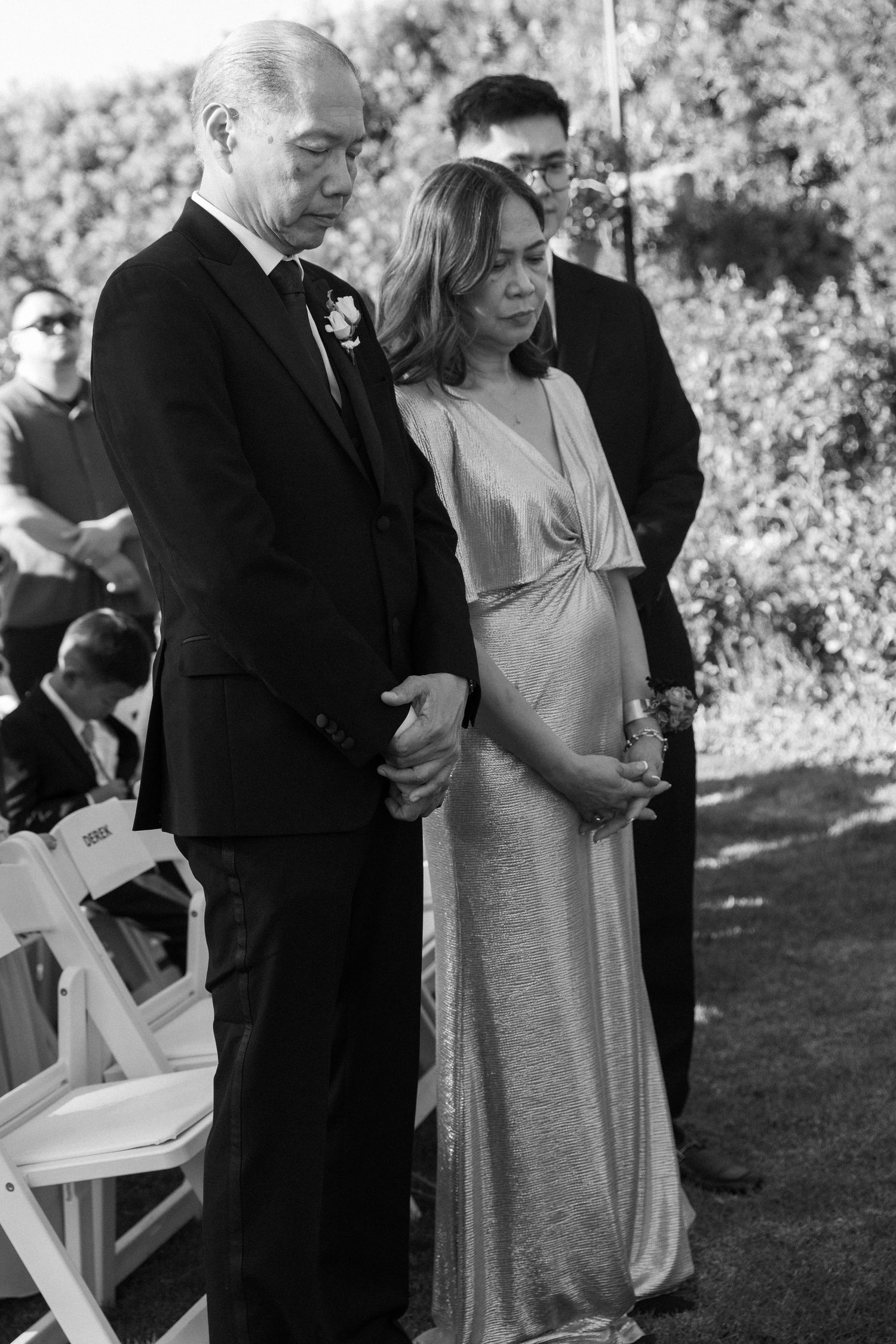 Wedding guests in formal attire seated on white chairs during outdoor ceremony.